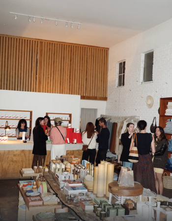 Inside a bustling modern boutique with white walls and wood accents, people browse clothing racks and rustic tables filled with candles, books, and home goods.