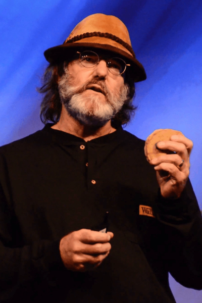 A man with a beard, glasses, and a brown hat holds a round food item and a pen, standing against a blue background.