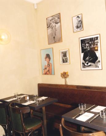 Restaurant interior with cream walls displaying framed photos, dark brown leather booth seating, and dark wooden tables set with glasses and cutlery.