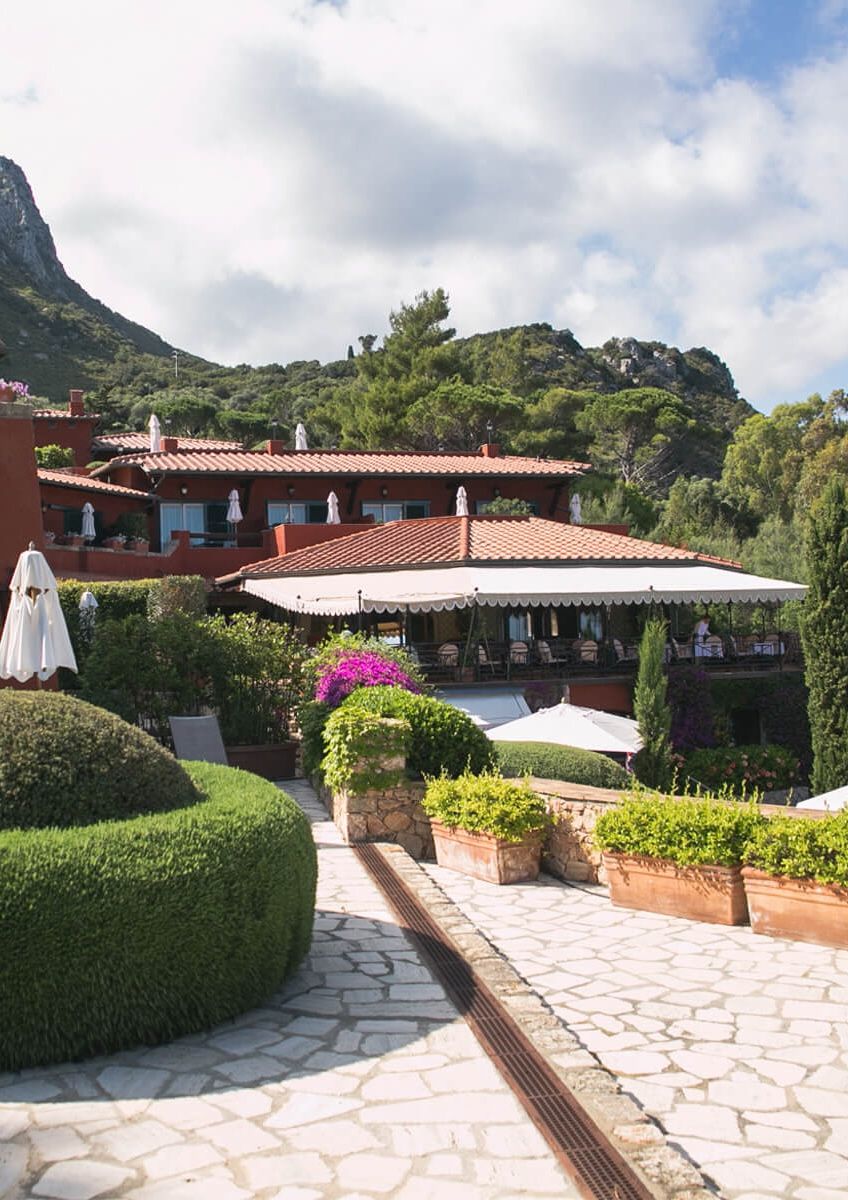 Mediterranean resort with red-roofed buildings, white umbrellas, lush gardens, and a stone path, nestled in green hills under a cloudy sky.