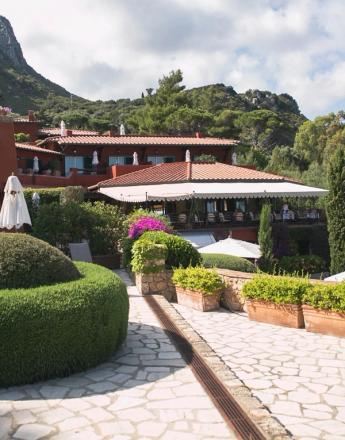 Mediterranean resort with red-roofed buildings, white umbrellas, lush gardens, and a stone path, nestled in green hills under a cloudy sky.
