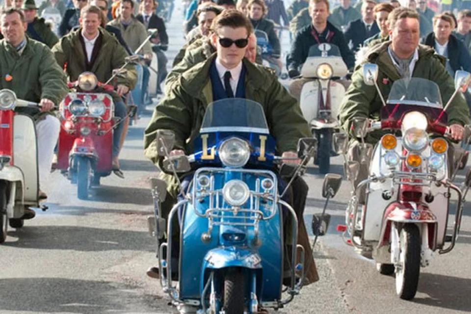 Homem usando parka por cima do terno andando de Vespa