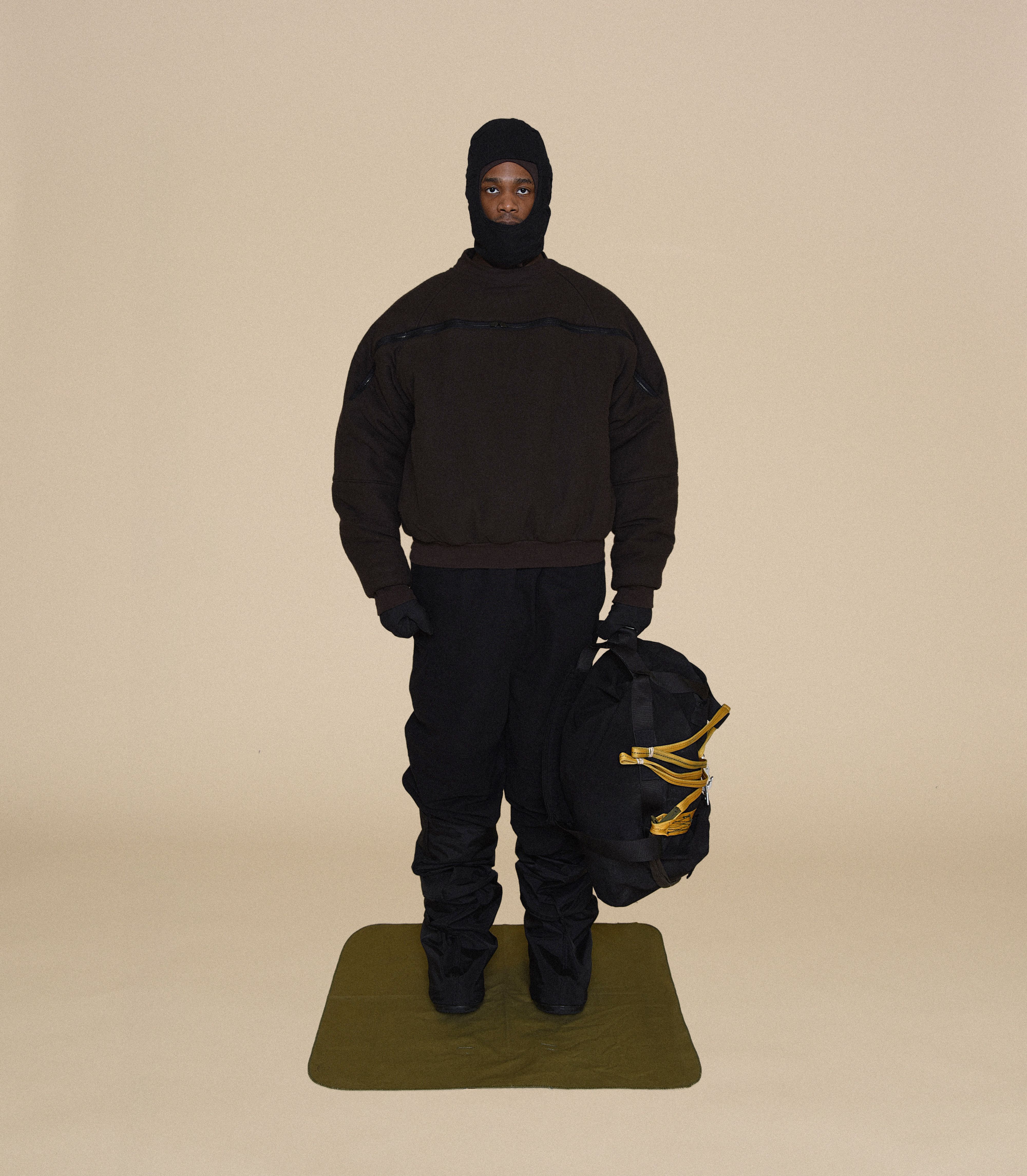 Man wearing HELMET LINER CAP, ANTI-EXPOSURE JACKET, SNIPER GLOVES and DUFFLE BAG From Bryan Jimenez fall/winter 2023.