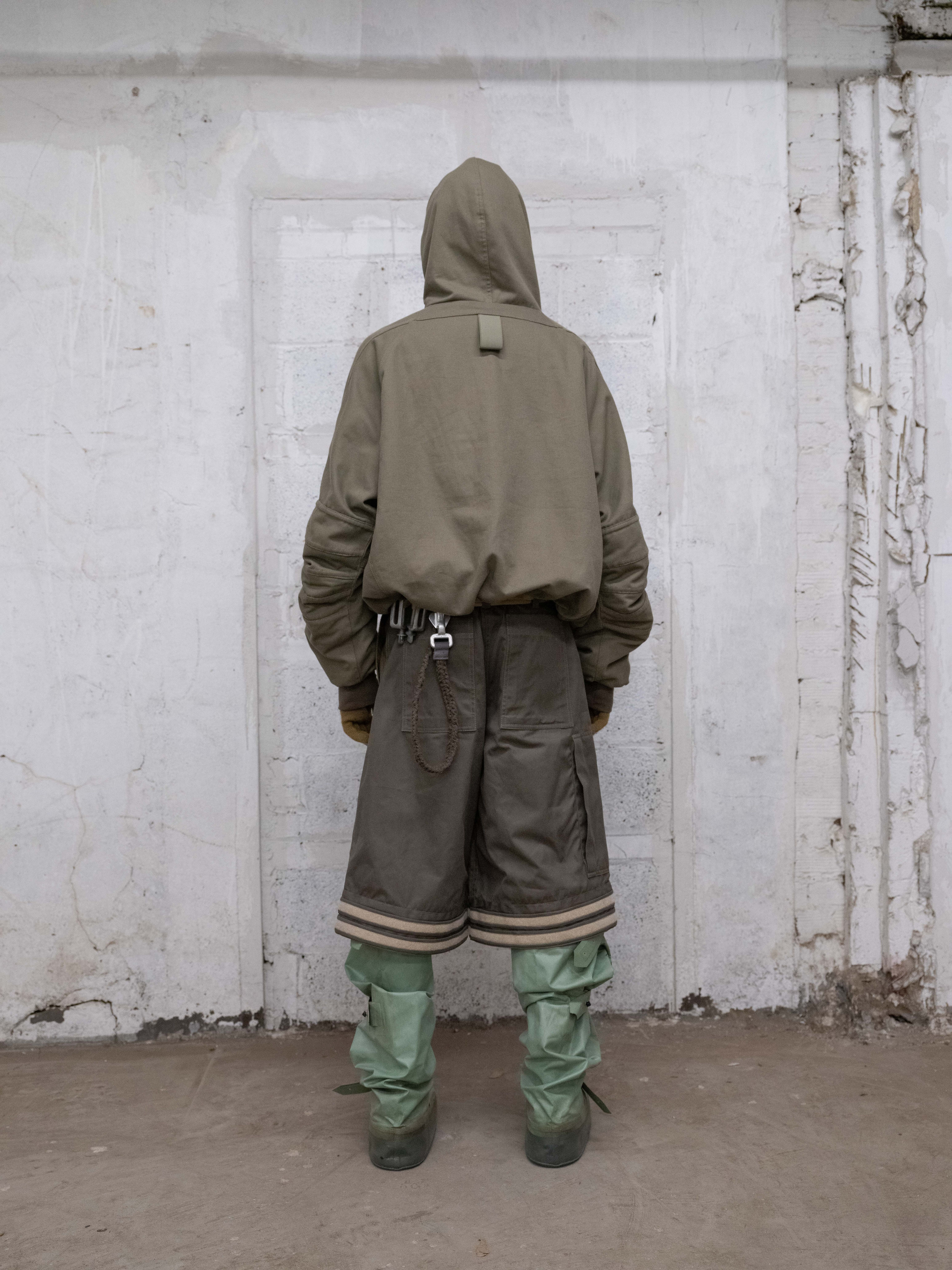 back view of man wearing mk olive jacket, bandolier belt with metal objects, cover boots and air bermudas from bryan jimenez fall/winter 2022