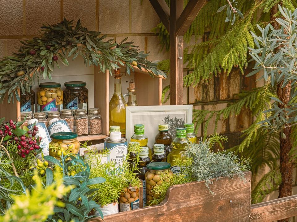 Esposizione di bottiglie di olio d'oliva e vasetti di olive tra rami d'ulivo e piante aromatiche