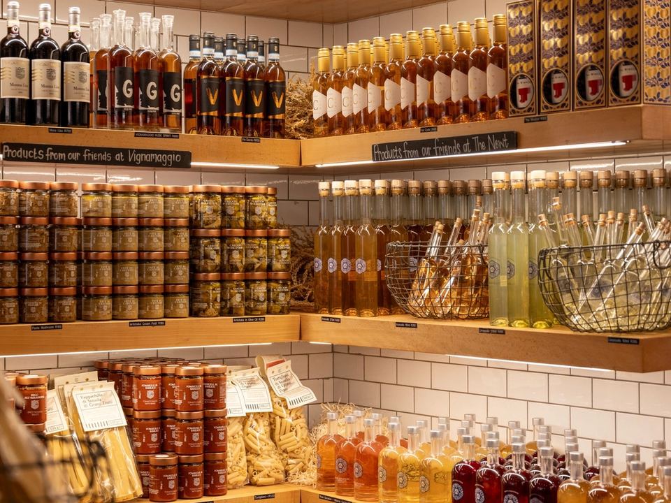 Scaffali di una bottega con bottiglie di olio e liquori, vasetti di conserve e confezioni di pasta etichettate