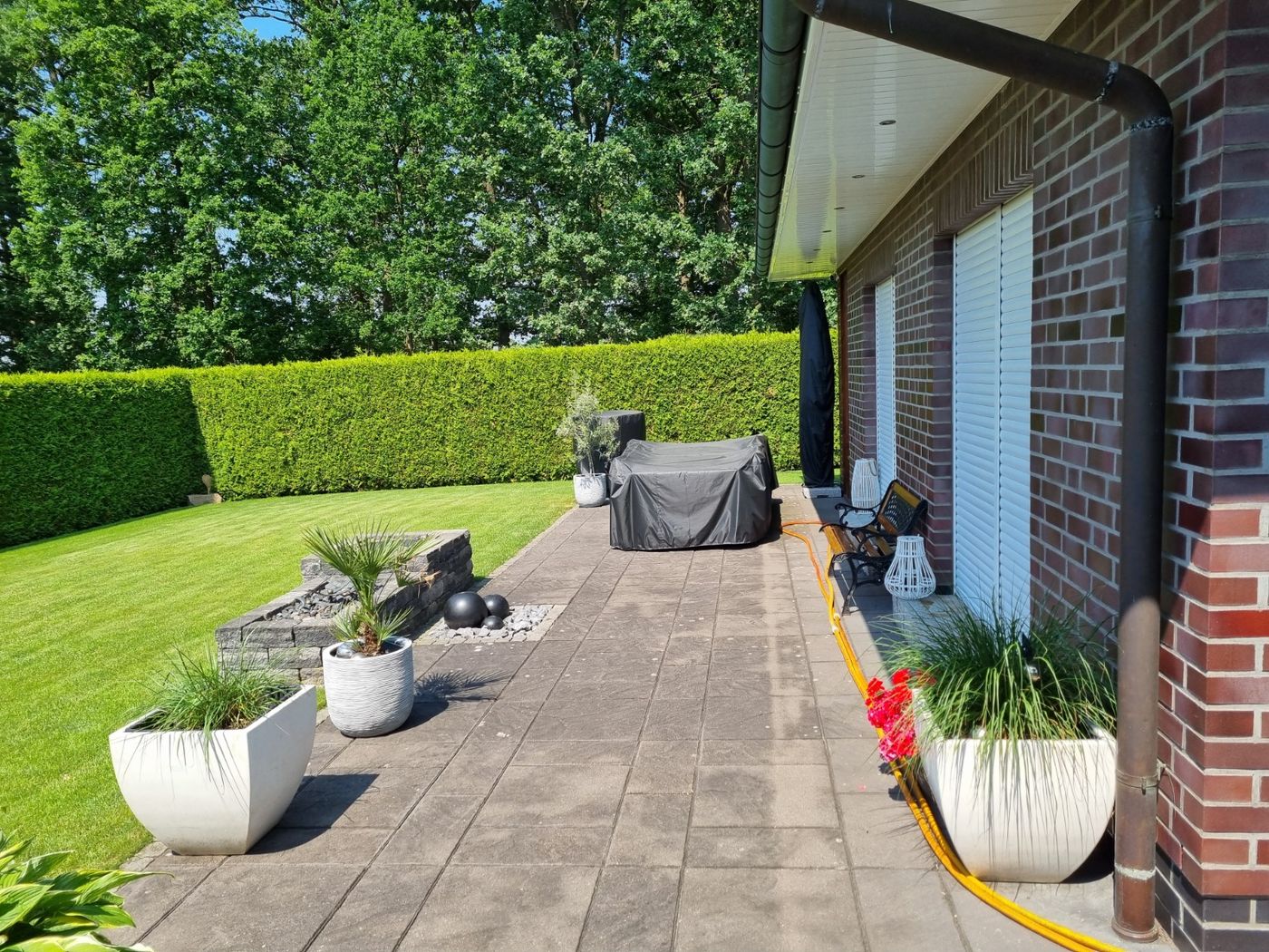 Seitlicher Blick auf die gepflasterte Terrasse mit Garten und Hausfassade vor dem Bau der Überdachung.