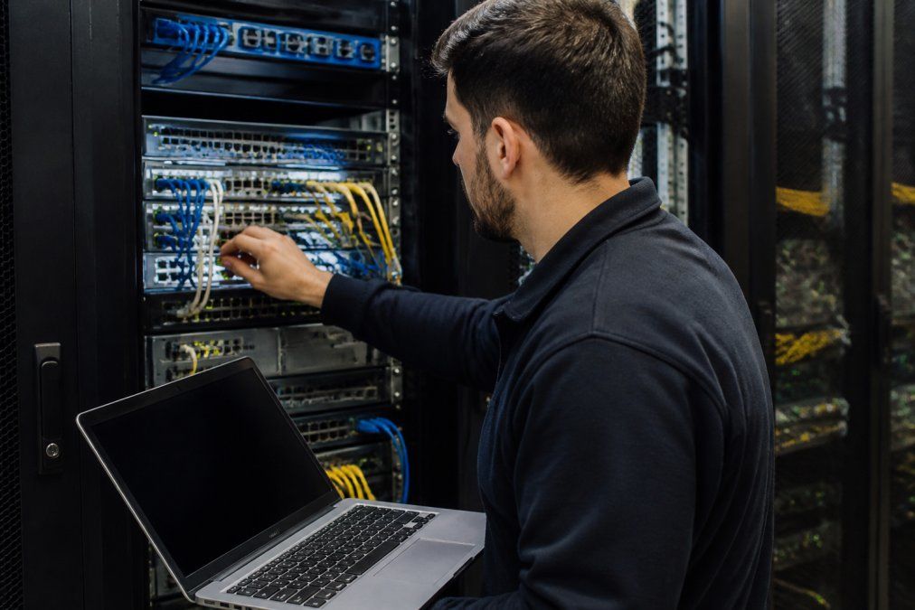 An IT technician in a server room checking network switches and patch cables, with a laptop showing network monitoring software