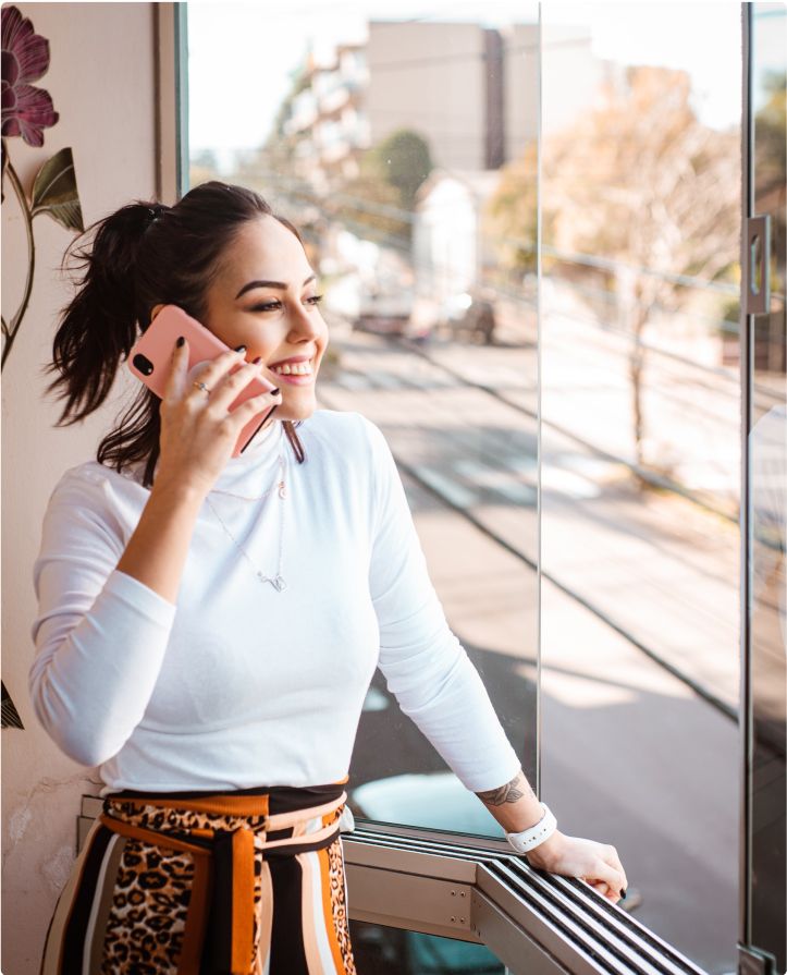 A young businesswoman smiles while talking on the cell phone while looking out a window onto a main street, leaning her left hand on a counter ledge.
