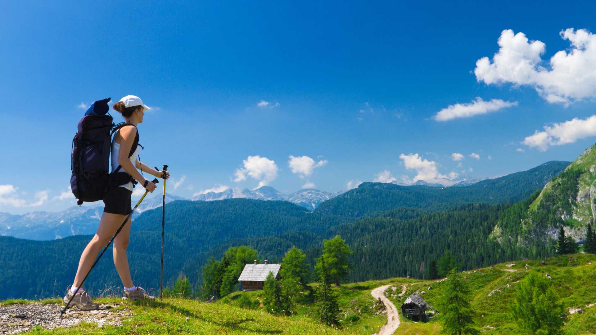 A person with a large backpack hikes with trekking poles on a grassy mountain trail under a blue sky with scattered clouds. A small wooden cabin is visible in the distance surrounded by trees and mountains.