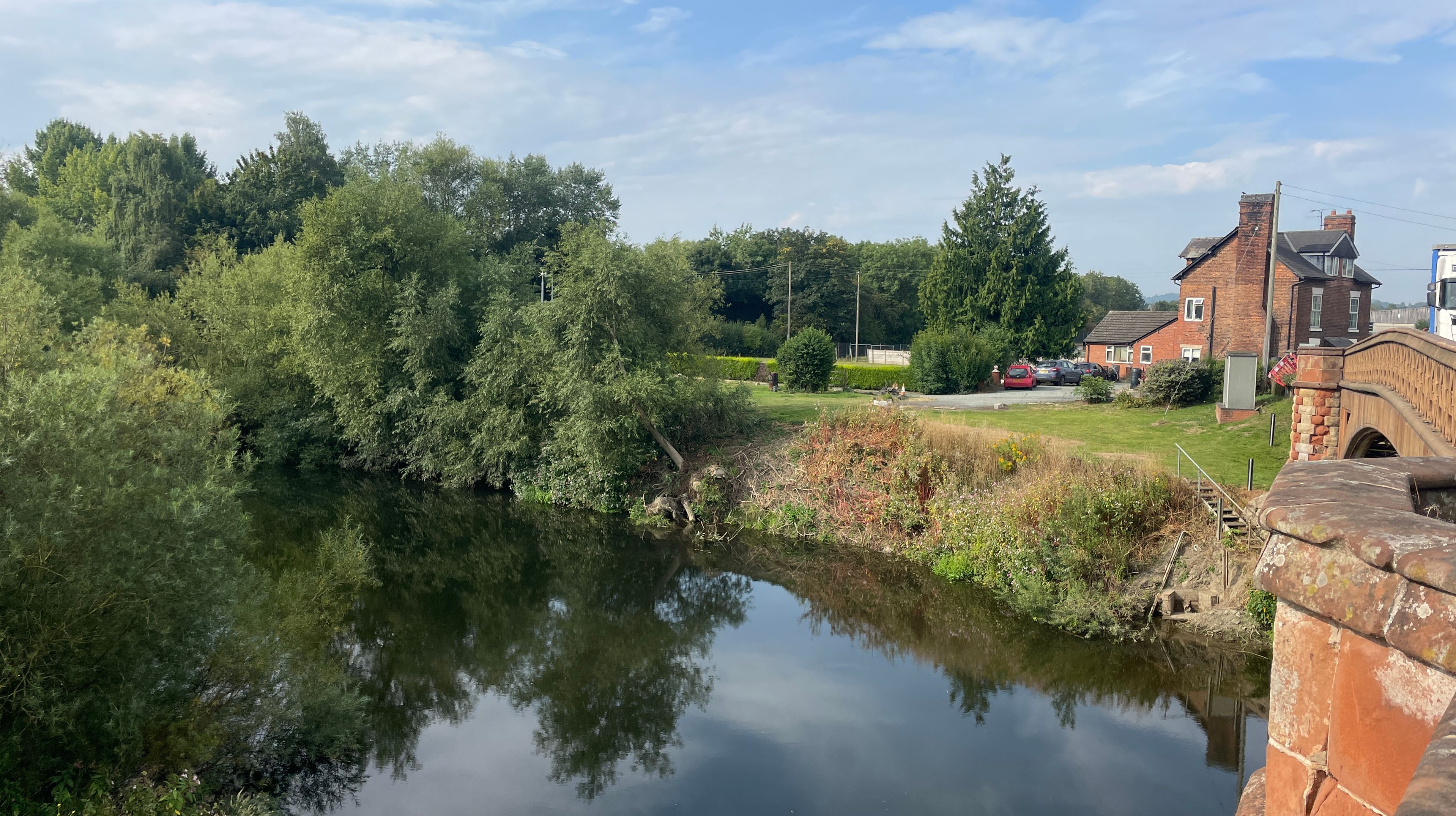 A wide river flows past lush green trees and bushes, with a stone bridge and brick buildings visible on the right bank under a partly cloudy sky.