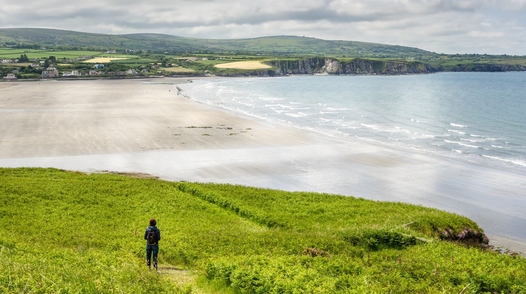 A lone hiker stands on a grassy hill overlooking a wide, sandy beach and the ocean under a cloudy sky.