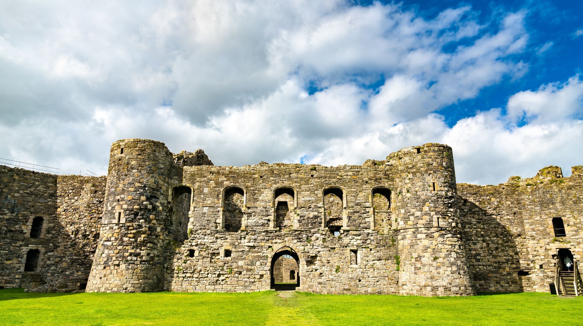 A stone castle with round towers and arched windows stands under a partly cloudy sky, with a green lawn in the foreground.