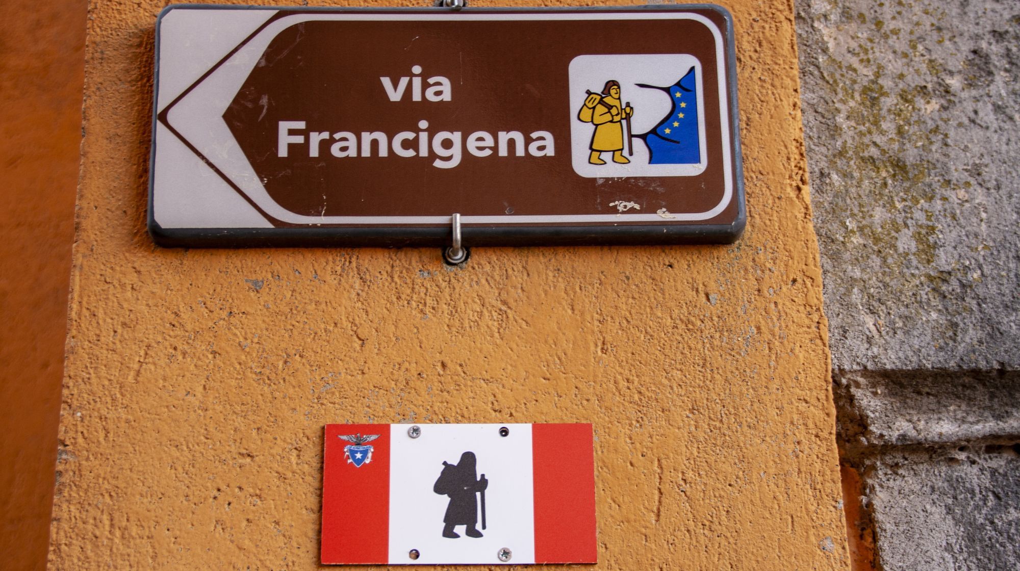 A brown sign with white text reading "via Francigena" and a symbol of a pilgrim, attached to an orange wall. Below it is a smaller rectangular sign with a red and white design and a black silhouette of a pilgrim.