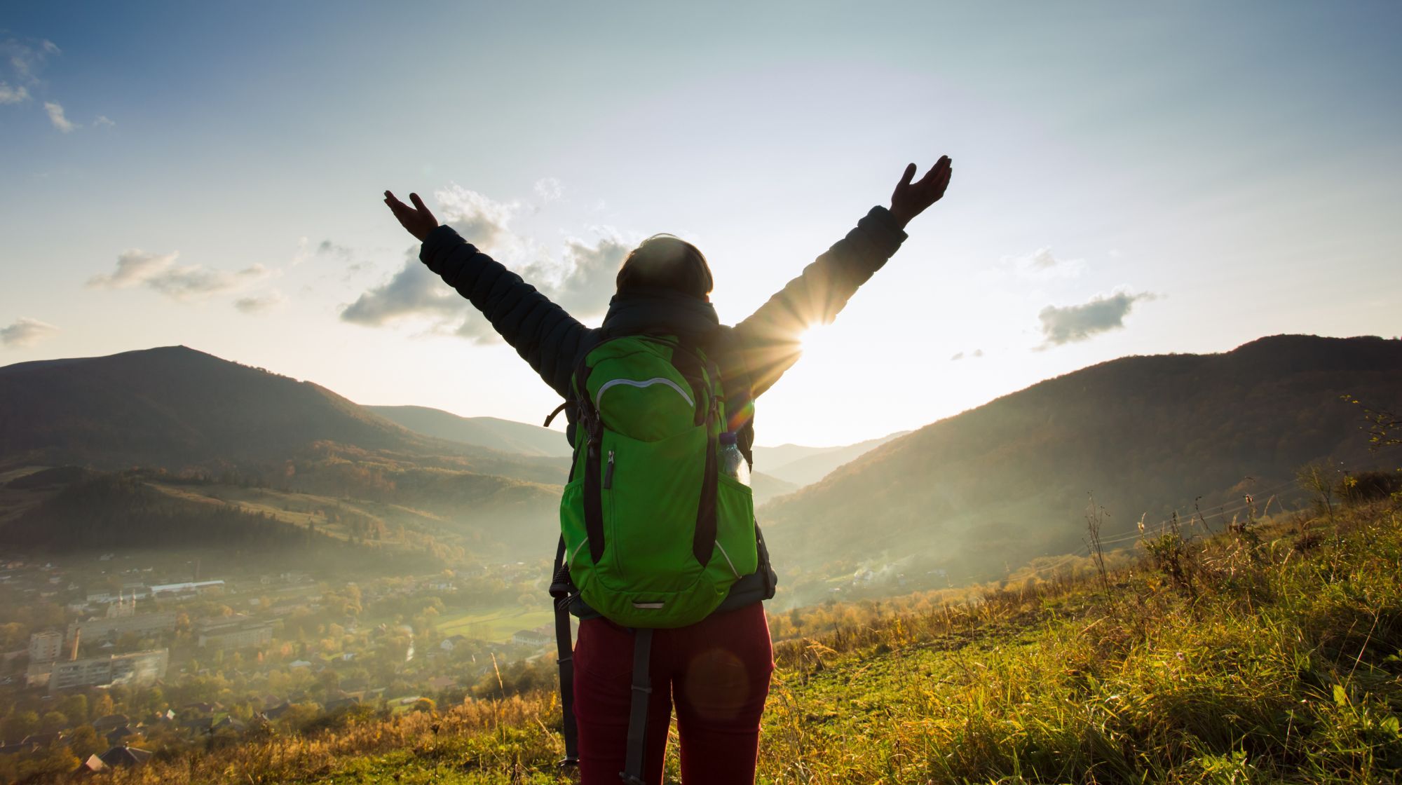 A person with a green backpack stands on a grassy hill with arms raised to the sky, silhouetted against the setting sun and mountains.