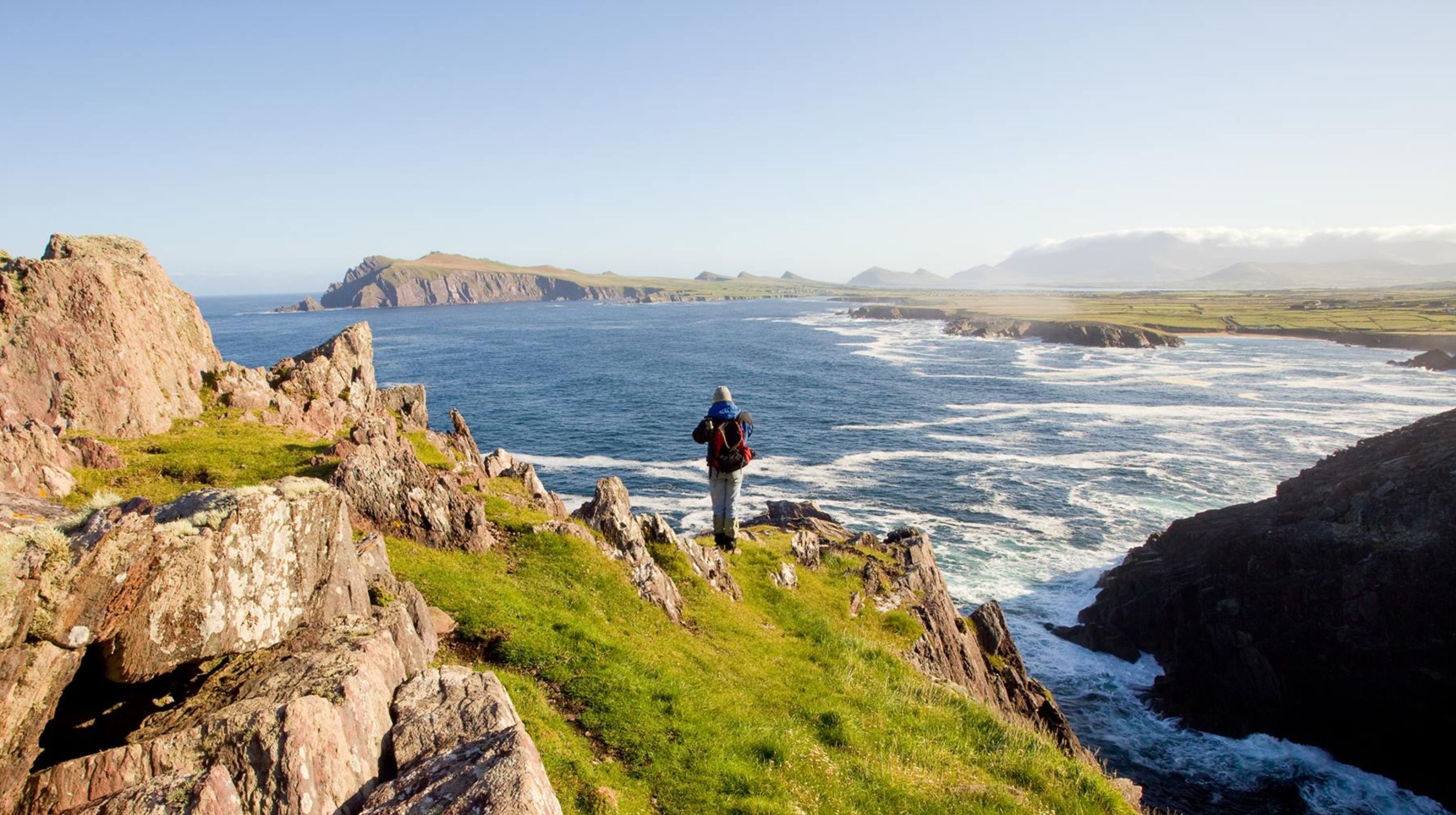 A person wearing a backpack stands on a grassy cliff overlooking the ocean with waves crashing against rocky shores.