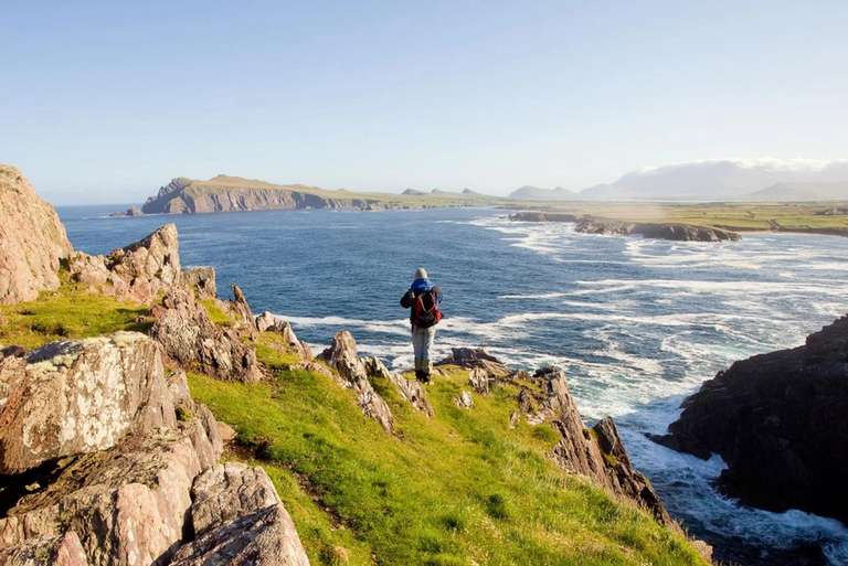 A person wearing a backpack stands on a grassy cliff overlooking the ocean with waves crashing against rocky shores.