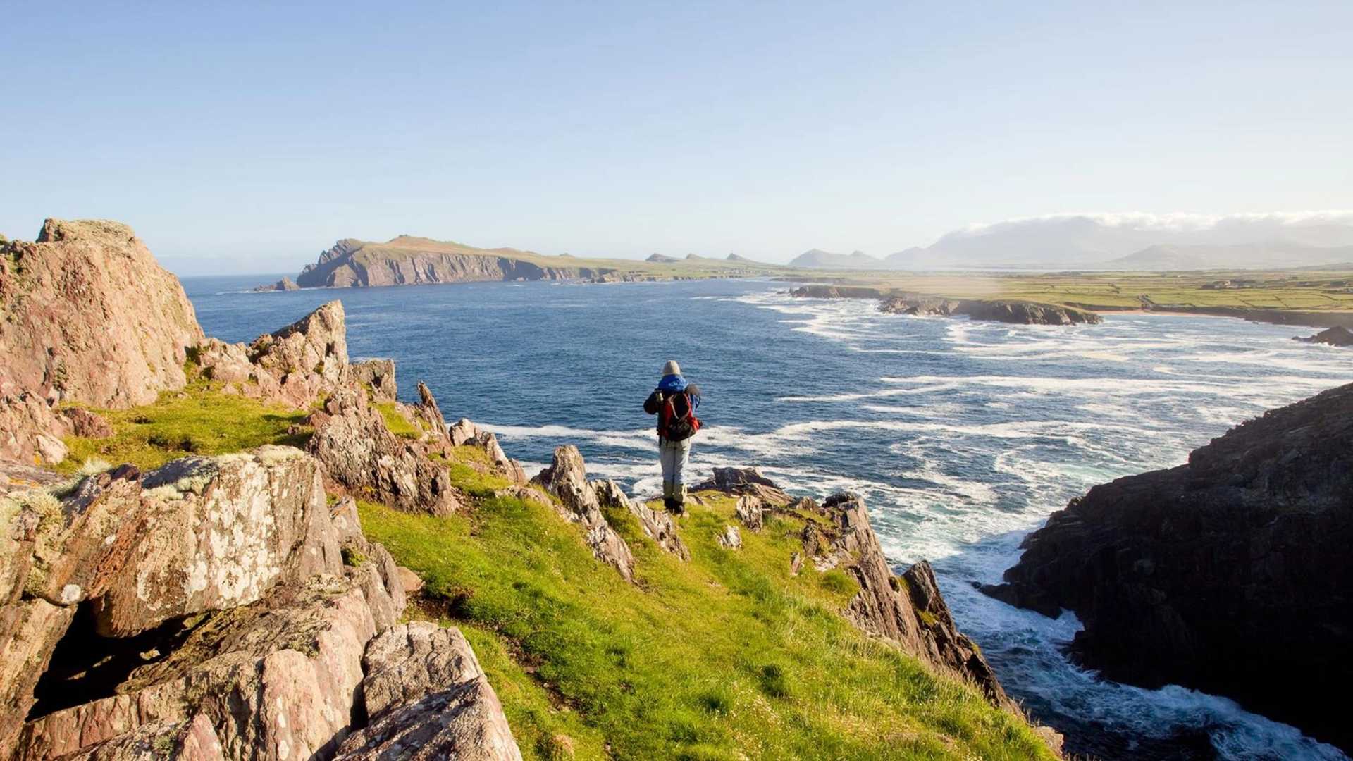 A person wearing a backpack stands on a grassy cliff overlooking the ocean with waves crashing against rocky shores.
