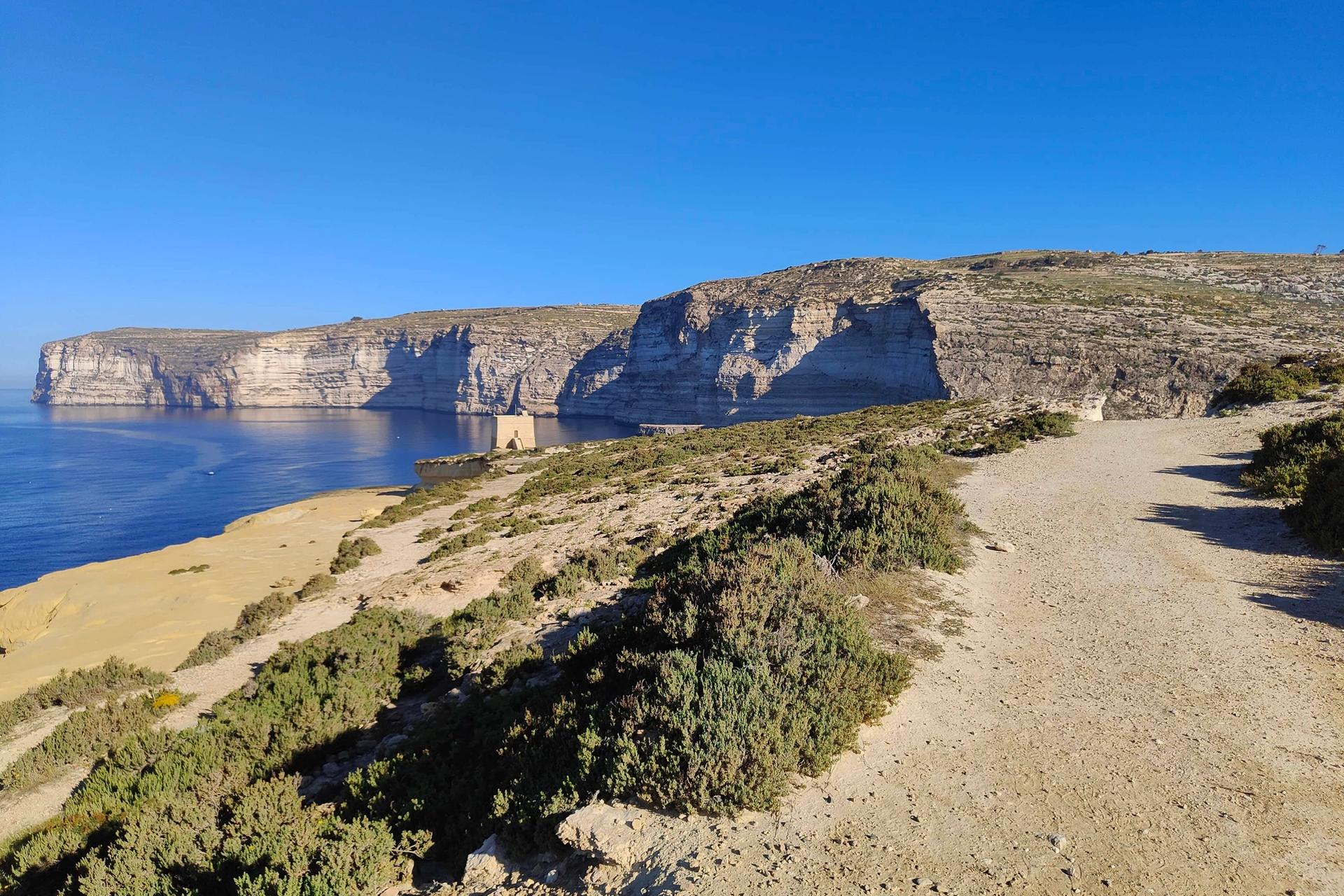 Dramatic limestone cliffs of Gozo with a coastal walking trail under clear blue skies.