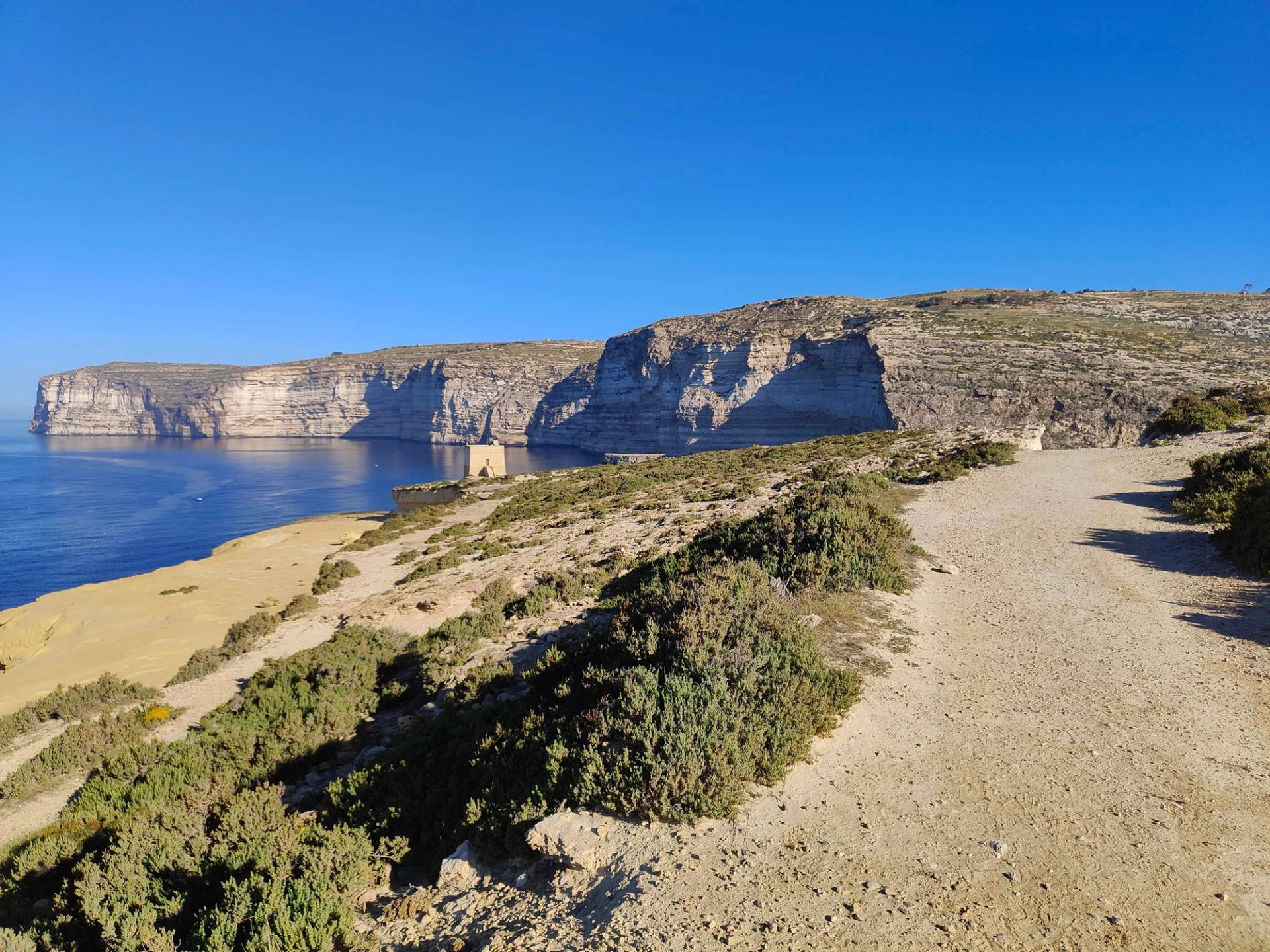 Dramatic limestone cliffs of Gozo with a coastal walking trail under clear blue skies.