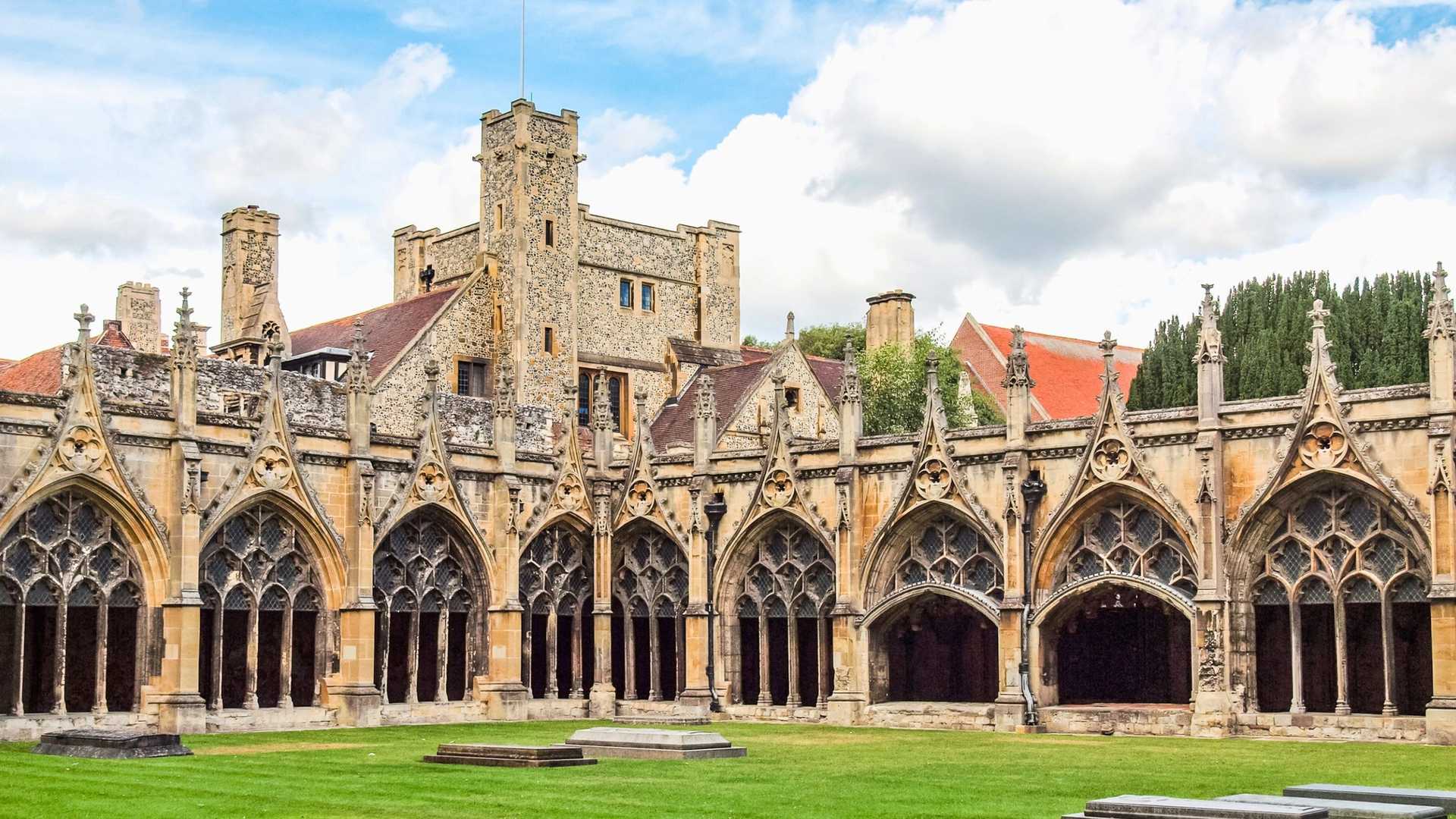 A cloister walk with arched windows and a green lawn in front of a stone building with a tower.