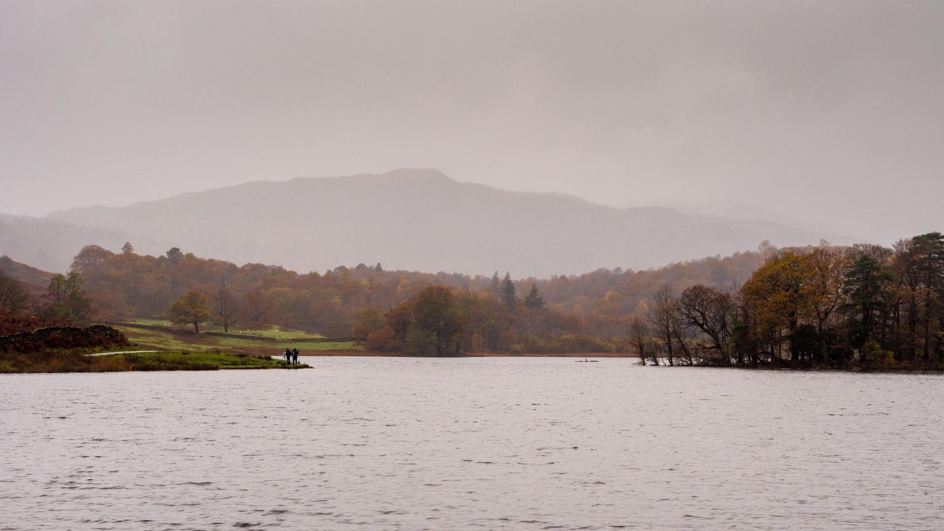 A wide view of a lake with forested shores and mountains in the distance under an overcast sky