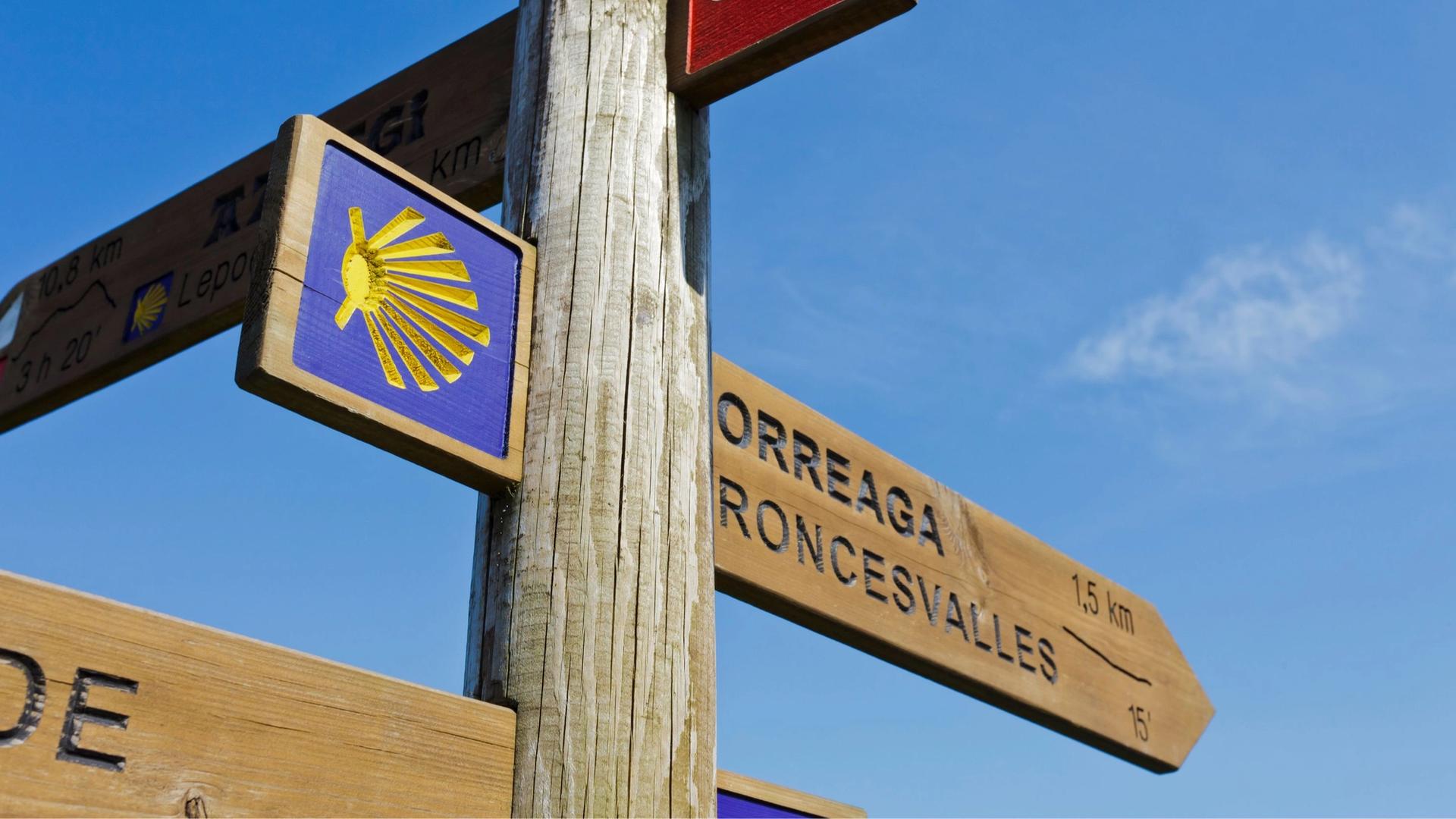 A wooden signpost with multiple directional arrows, including one with the yellow scallop shell symbol of the Camino de Santiago, against a clear blue sky.