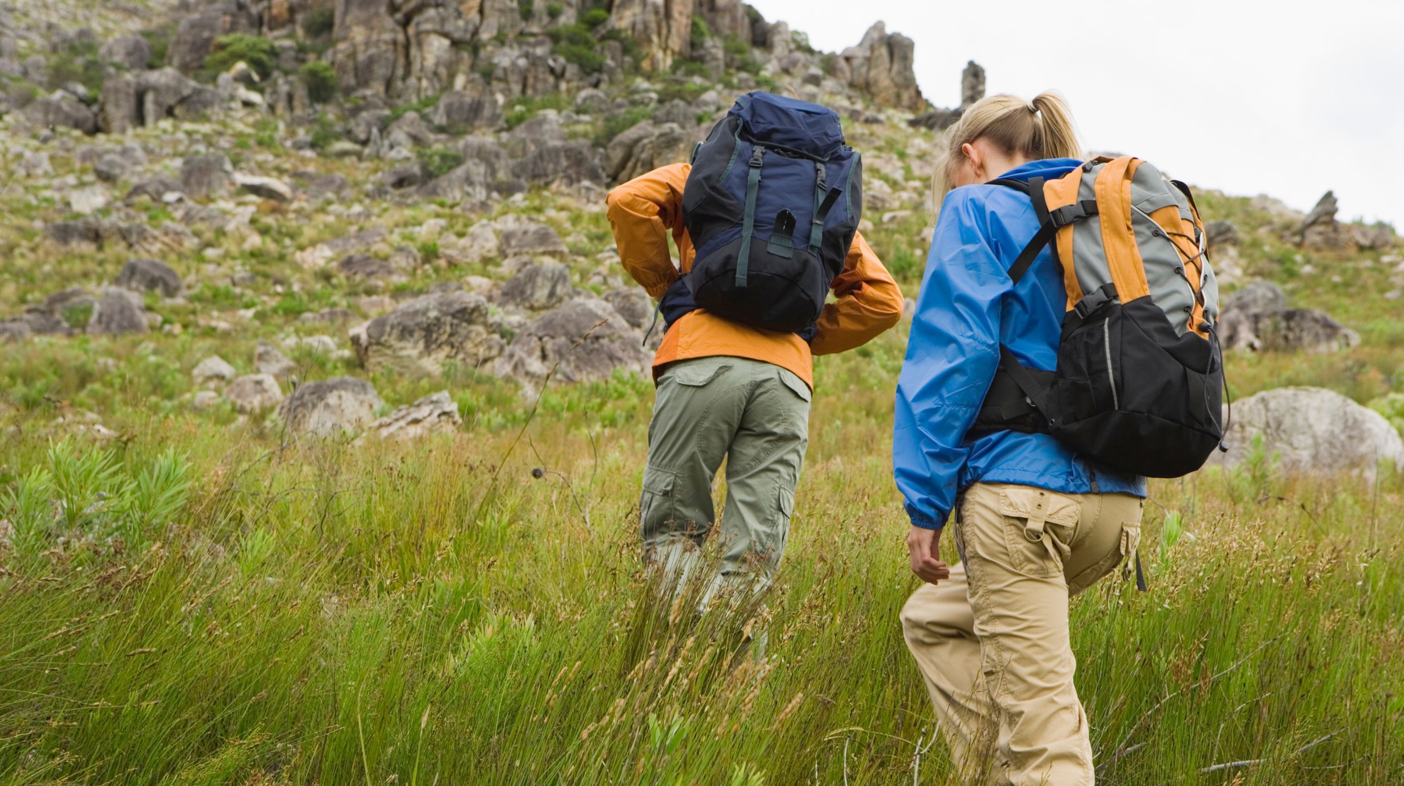 Two individuals with backpacks hiking through a grassy, rocky terrain.