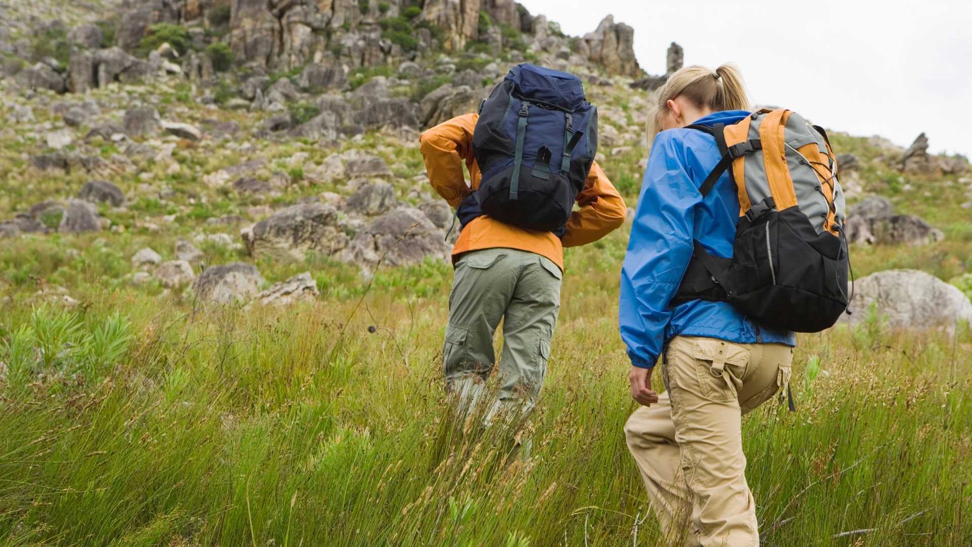 Two individuals with backpacks hiking through a grassy, rocky terrain.