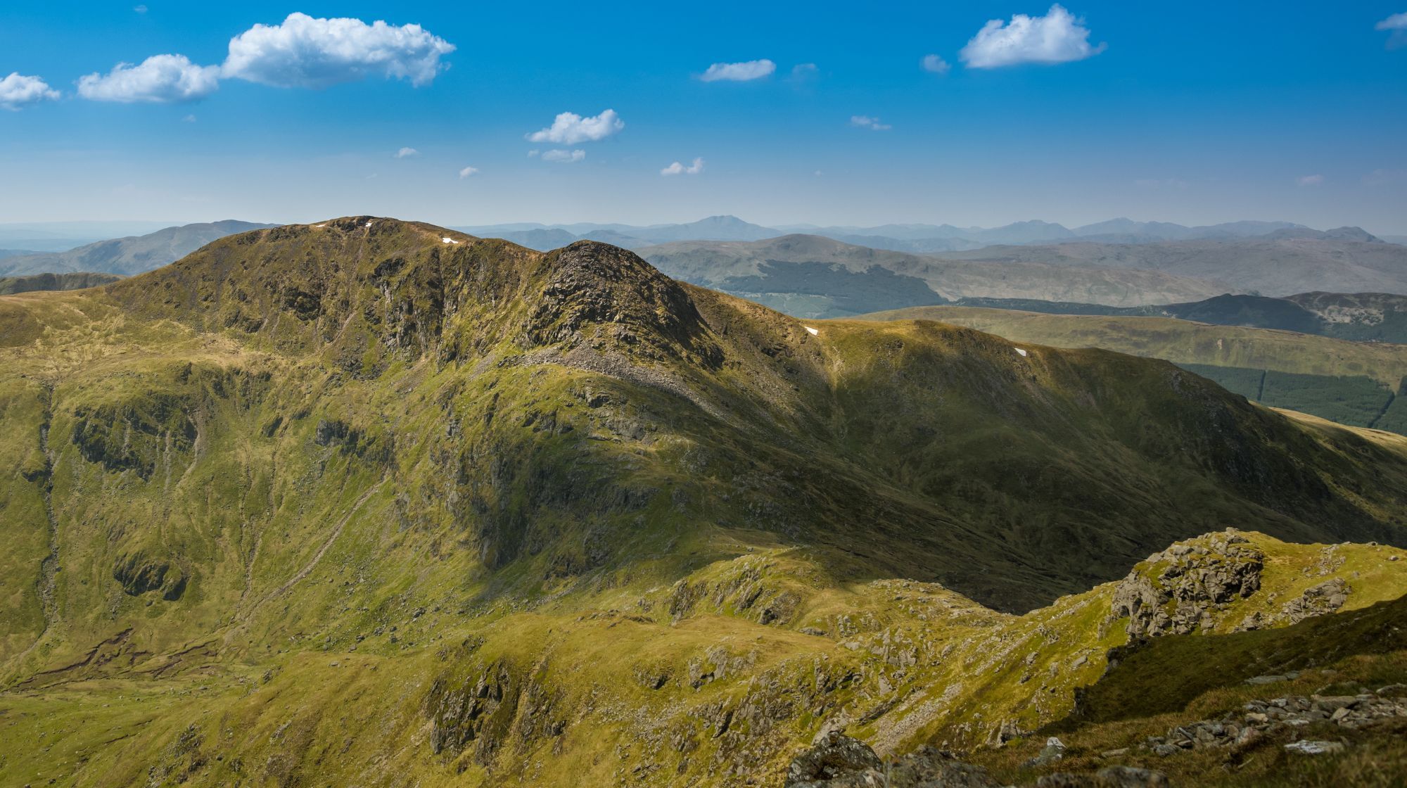 A wide panoramic view of rolling green hills and mountains under a bright blue sky with scattered white clouds.