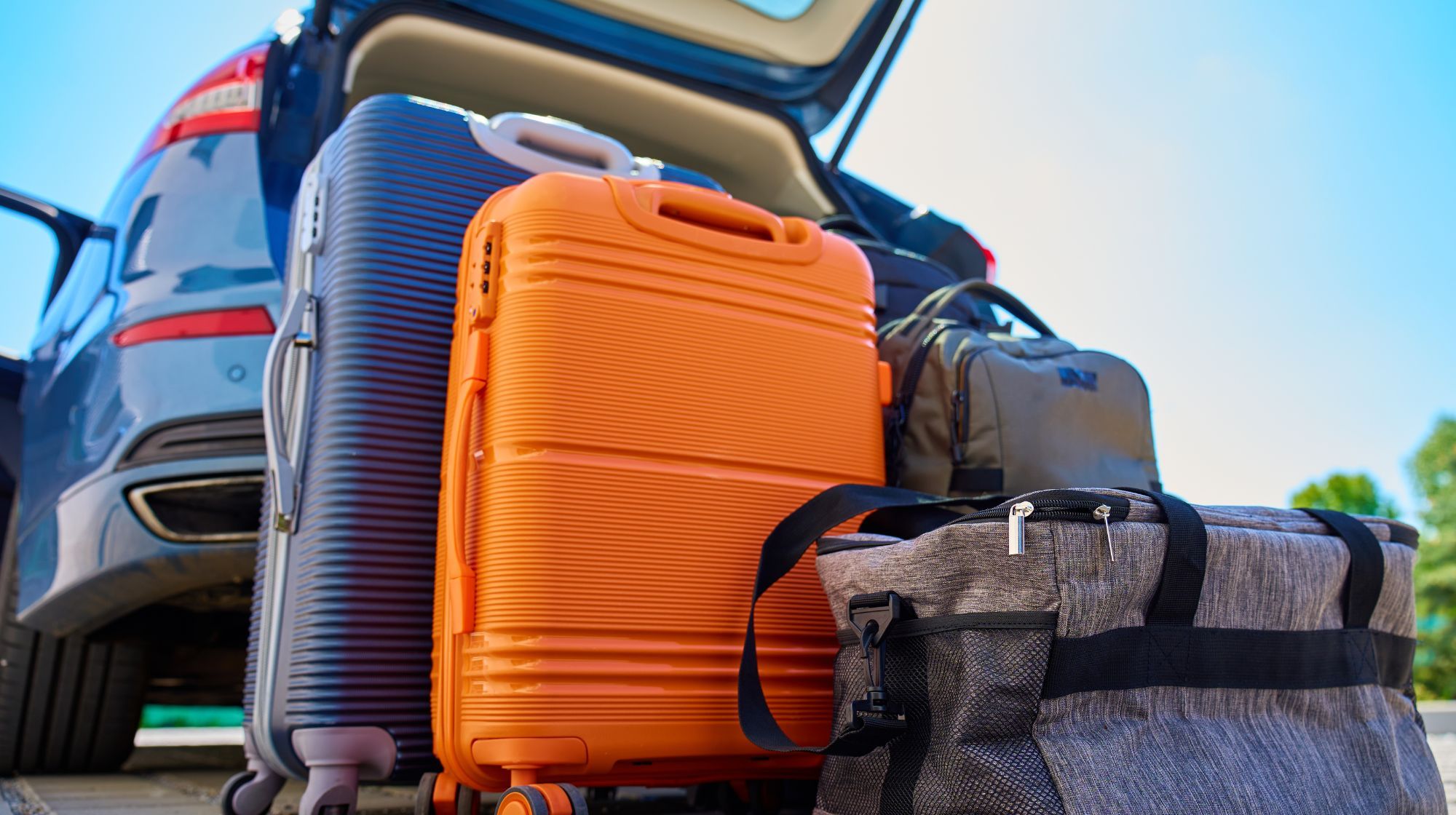 Several pieces of luggage are packed into the open trunk of a car, ready for a trip. The luggage includes a large orange suitcase, a dark blue ribbed suitcase, and two duffel bags.