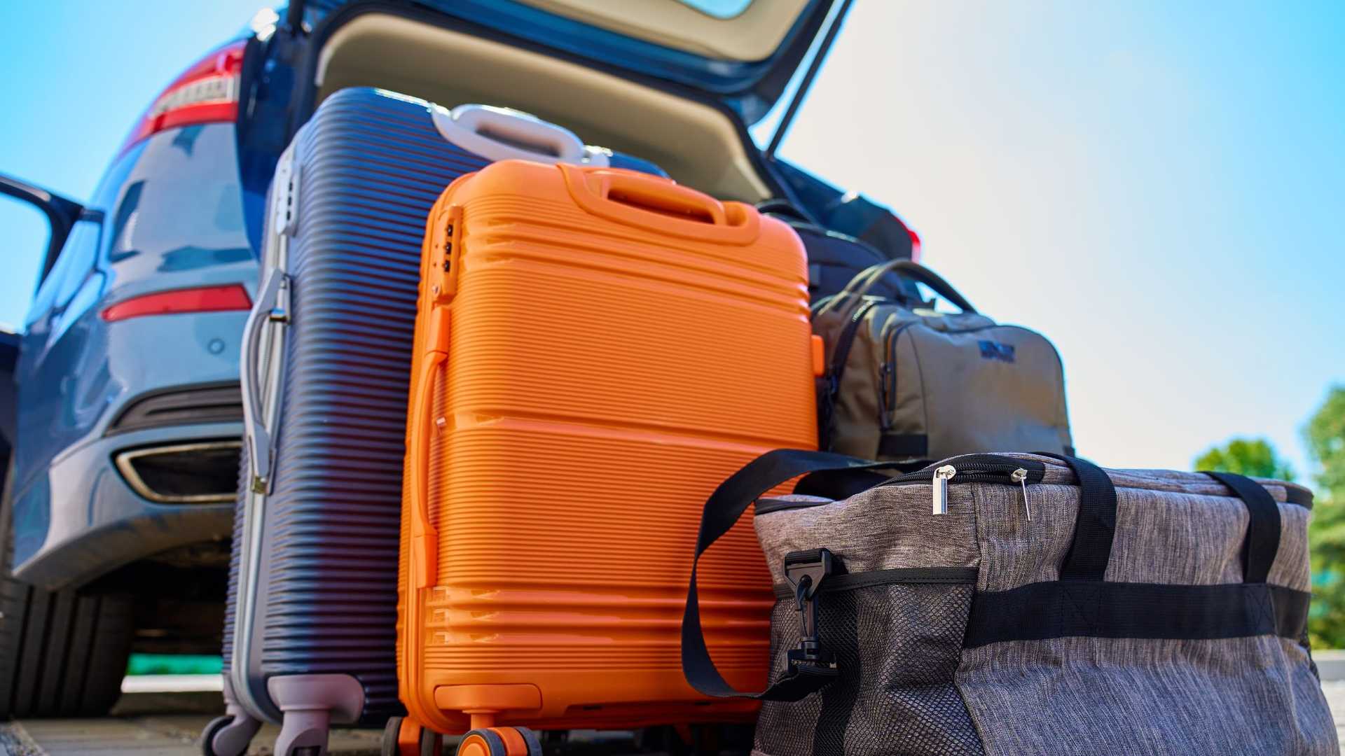 Several pieces of luggage are packed into the open trunk of a car, ready for a trip. The luggage includes a large orange suitcase, a dark blue ribbed suitcase, and two duffel bags.
