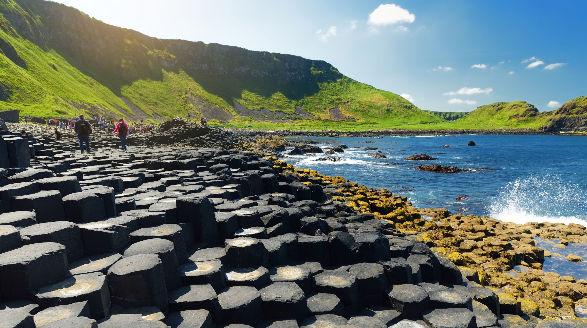 Hexagonal basalt columns form a unique landscape along the coast with green hills in the background and the blue sea on the right.