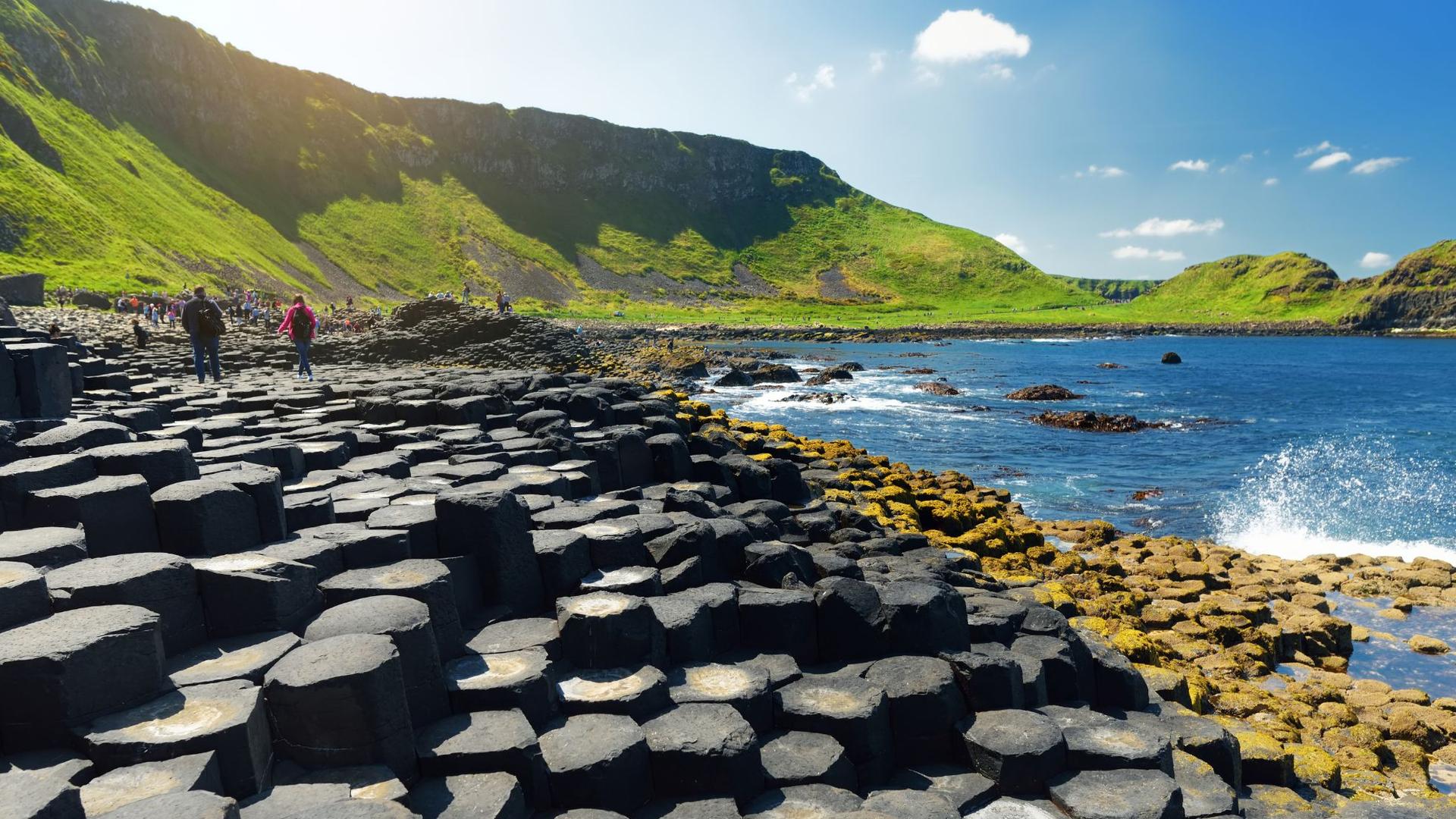 Hexagonal basalt columns form a unique landscape along the coast with green hills in the background and the blue sea on the right.