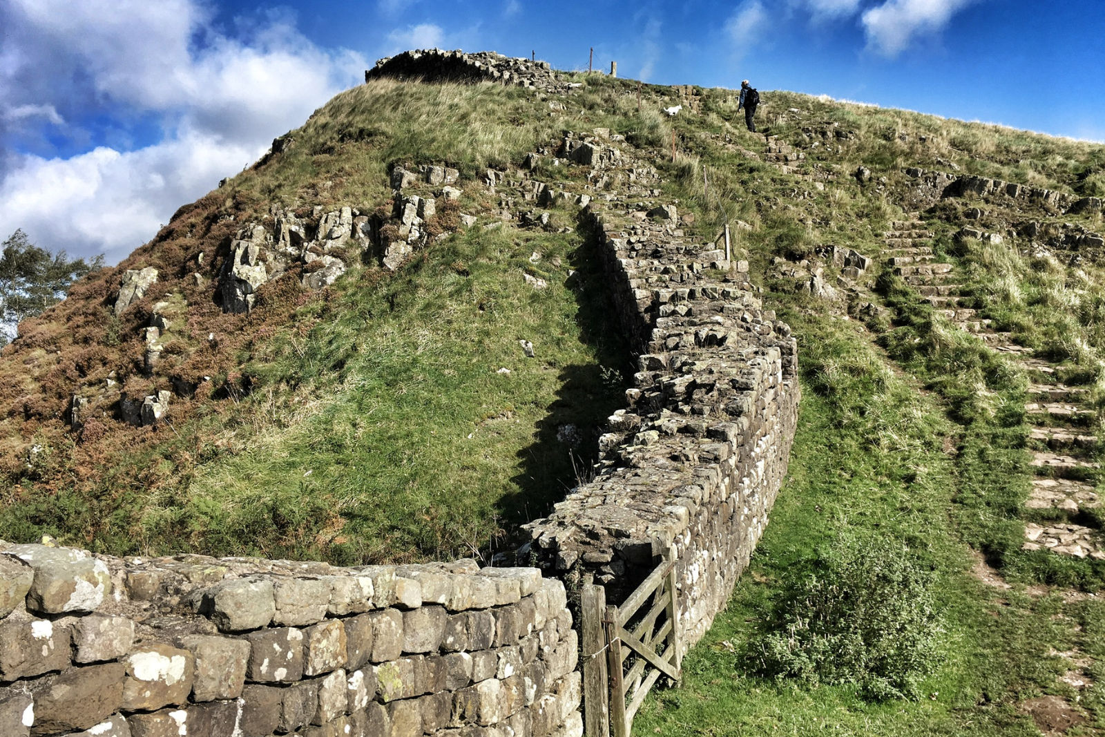Hadrian's Wall climbing a steep grassy ridge, with stone steps following the hillside.
