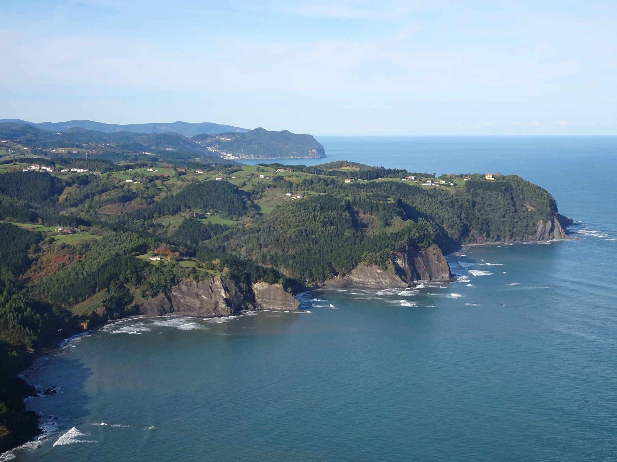 Panoramic view of the Basque Coast, with rugged forested cliffs, rolling green hills and scattered farmhouses overlooking the Bay of Biscay.