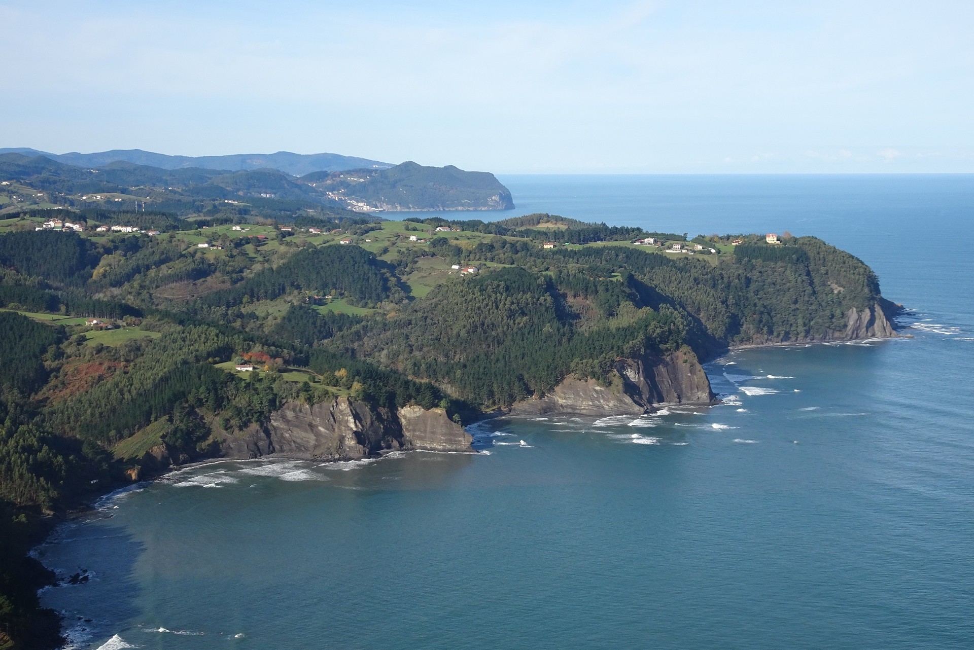Panoramic view of the Basque Coast, with rugged forested cliffs, rolling green hills and scattered farmhouses overlooking the Bay of Biscay.