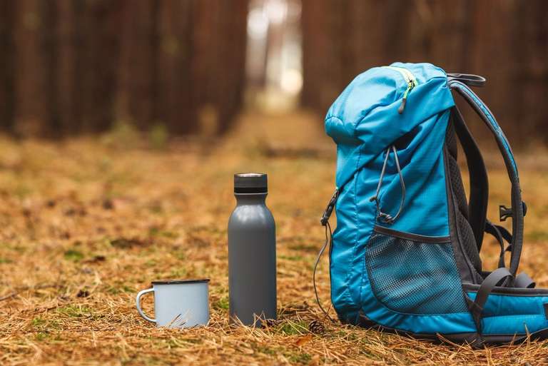 A blue backpack, a gray water bottle, and a light blue enamel mug sit on the ground in a forest setting.