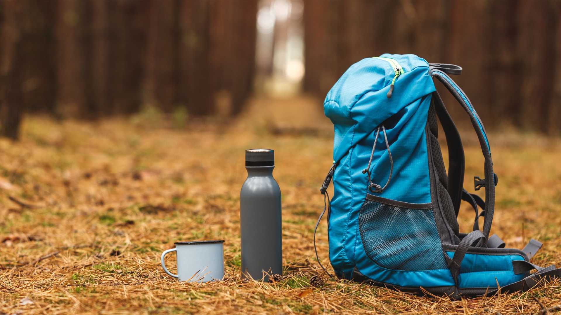 A blue backpack, a gray water bottle, and a light blue enamel mug sit on the ground in a forest setting.