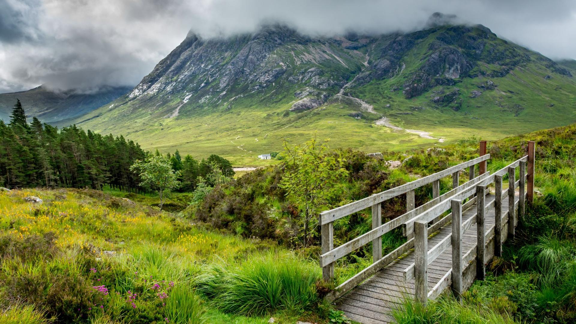 A wooden footbridge leads to a green valley and mountain partially covered in clouds
