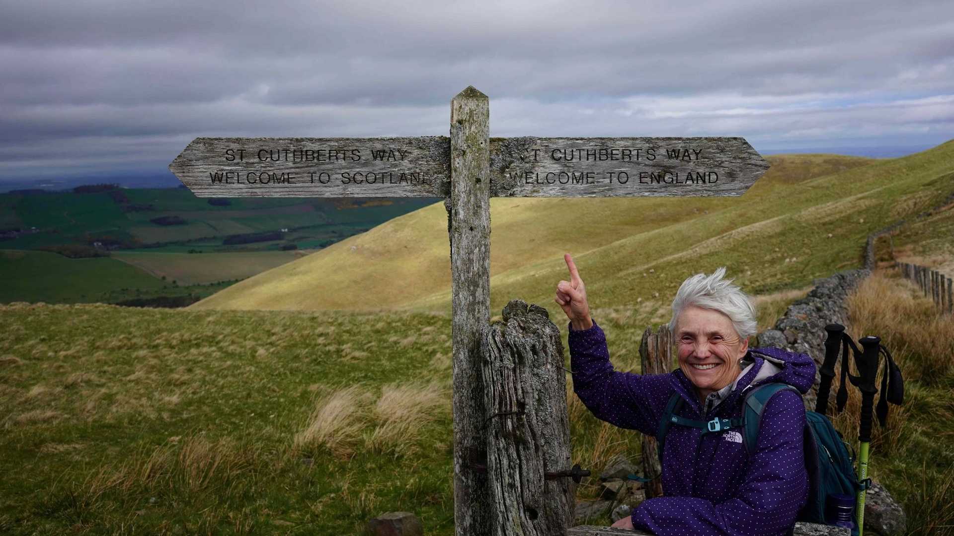 A person points at a wooden signpost marking the St. Cuthbert's Way, with one arrow pointing to Scotland and the other to England.
