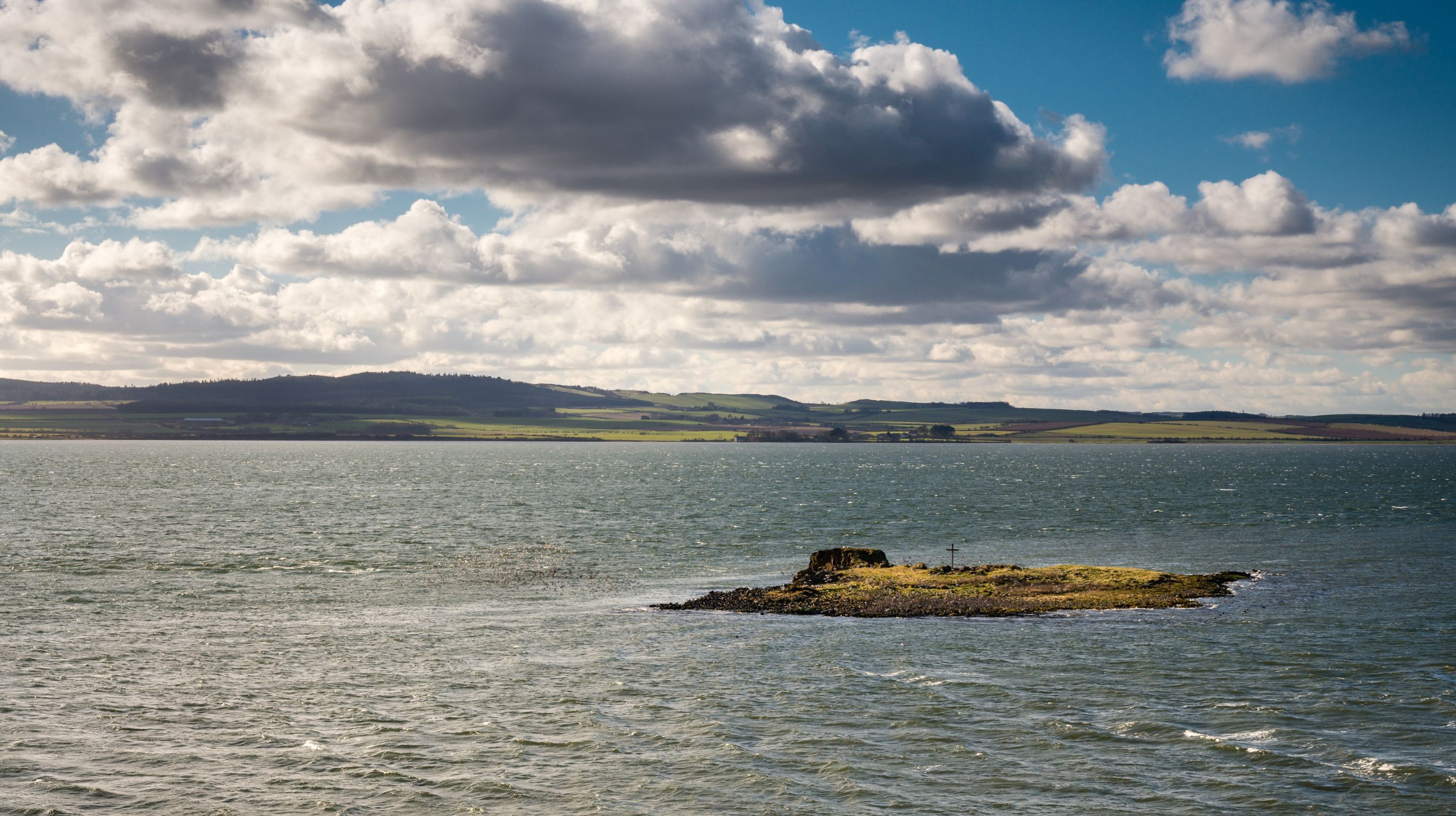 A small, rocky island covered in grass sits in a body of water under a cloudy sky with a distant shoreline.