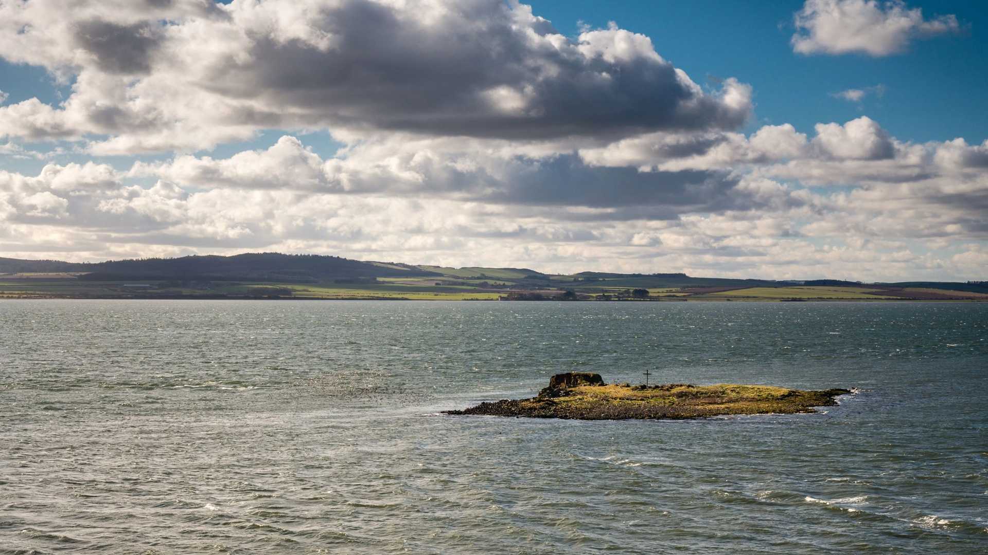 A small, rocky island covered in grass sits in a body of water under a cloudy sky with a distant shoreline.