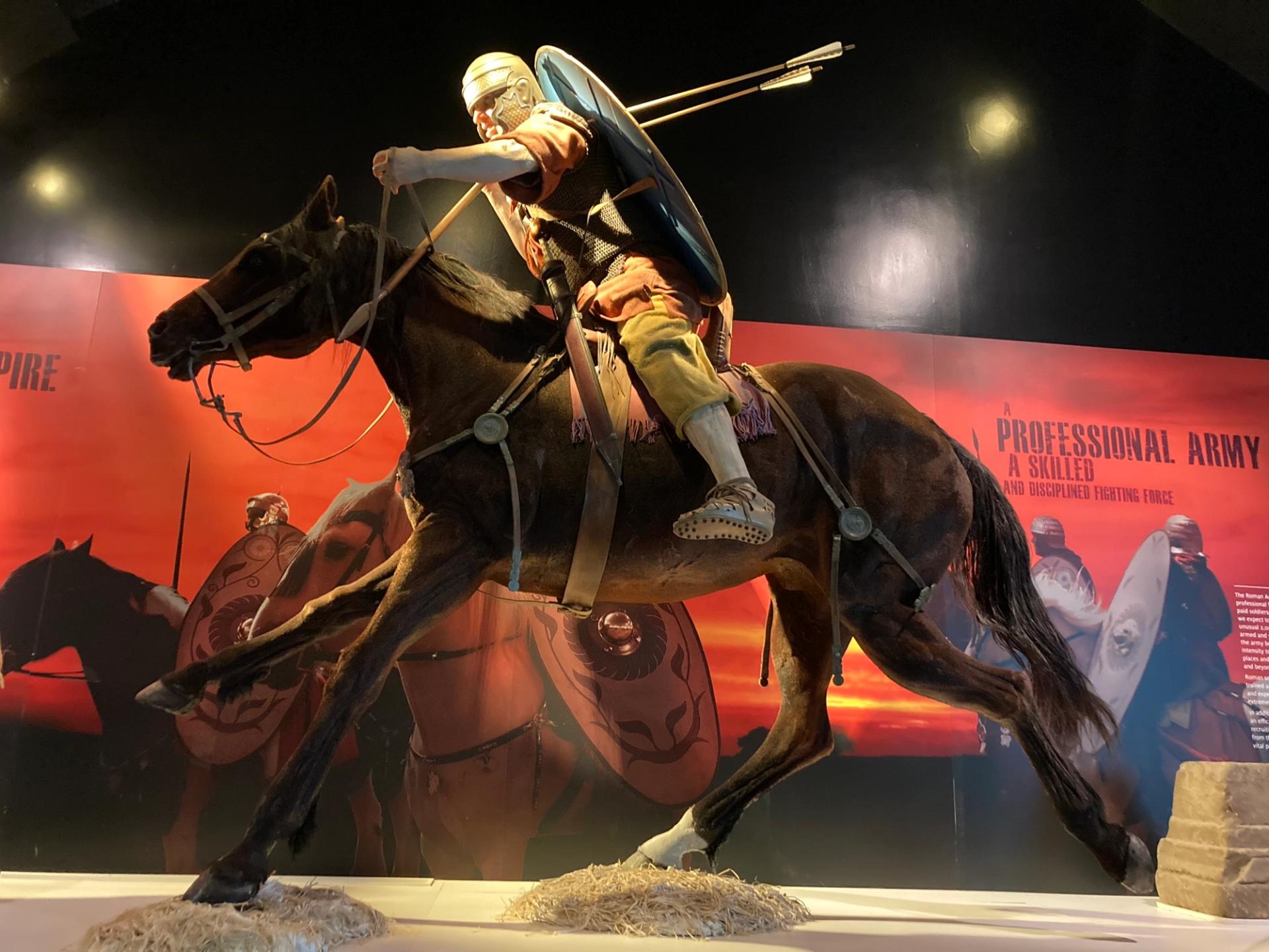 A realistic display of a Roman soldier on horseback, wearing armor and holding a shield and spear, with a backdrop depicting other soldiers and text about a professional army.
