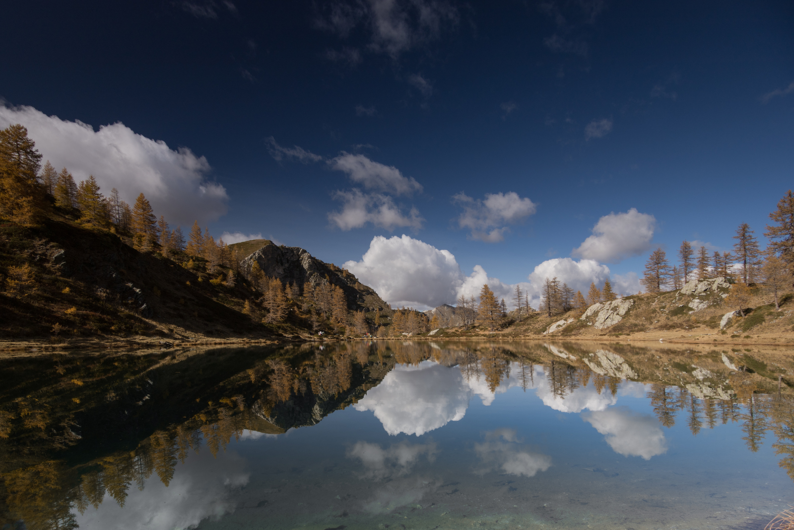 Still waters of Lago Nero reflecting larch trees, clouds and the surrounding mountains of Val Maira.