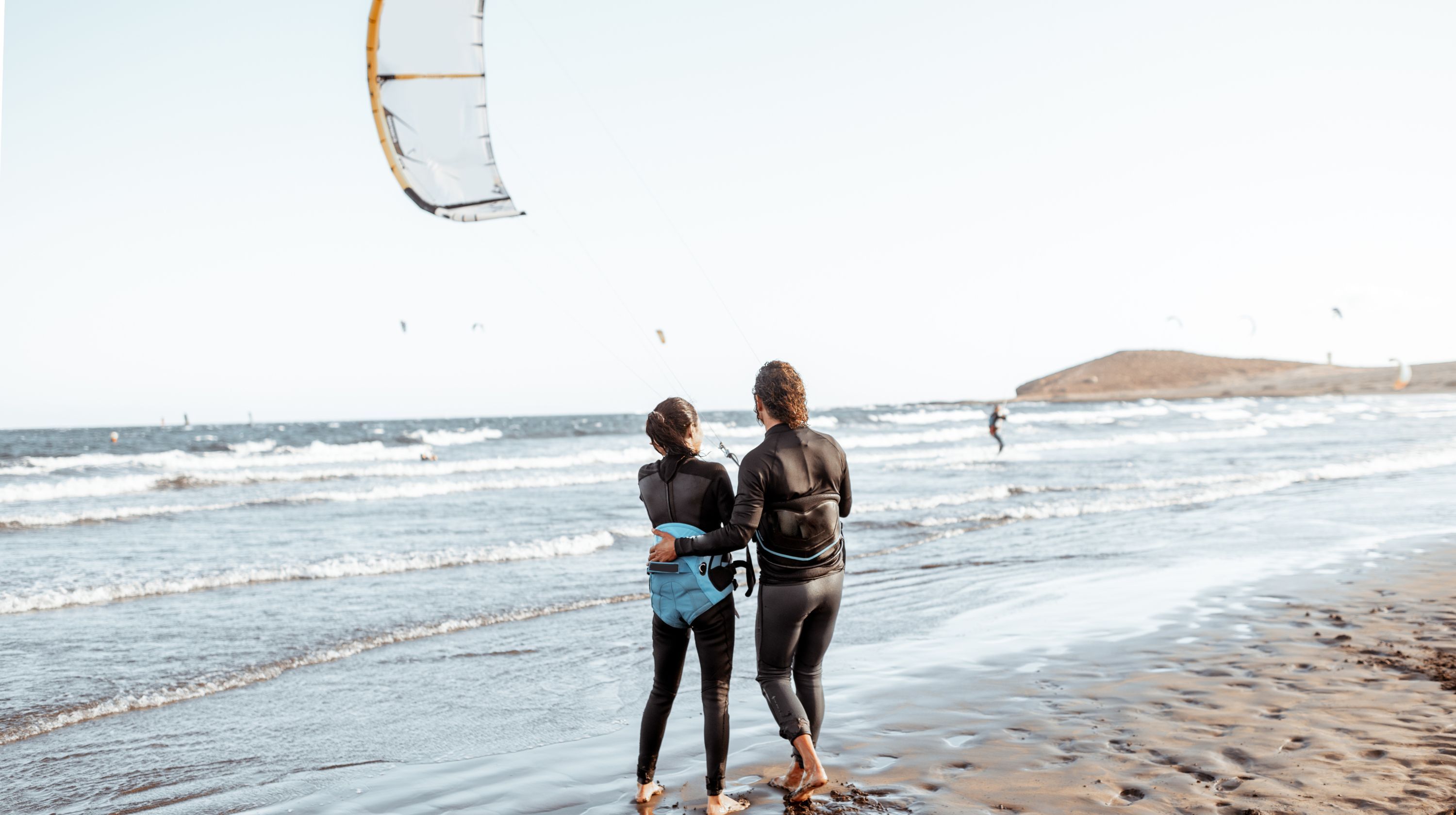 Two people in wetsuits stand on a beach looking out at the ocean where a kiteboarder is in the water and a kite is in the air.