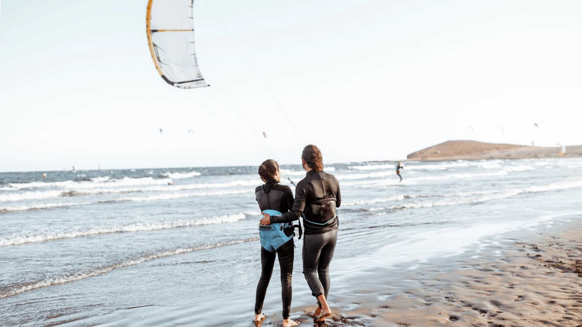 Two people in wetsuits stand on a beach looking out at the ocean where a kiteboarder is in the water and a kite is in the air.