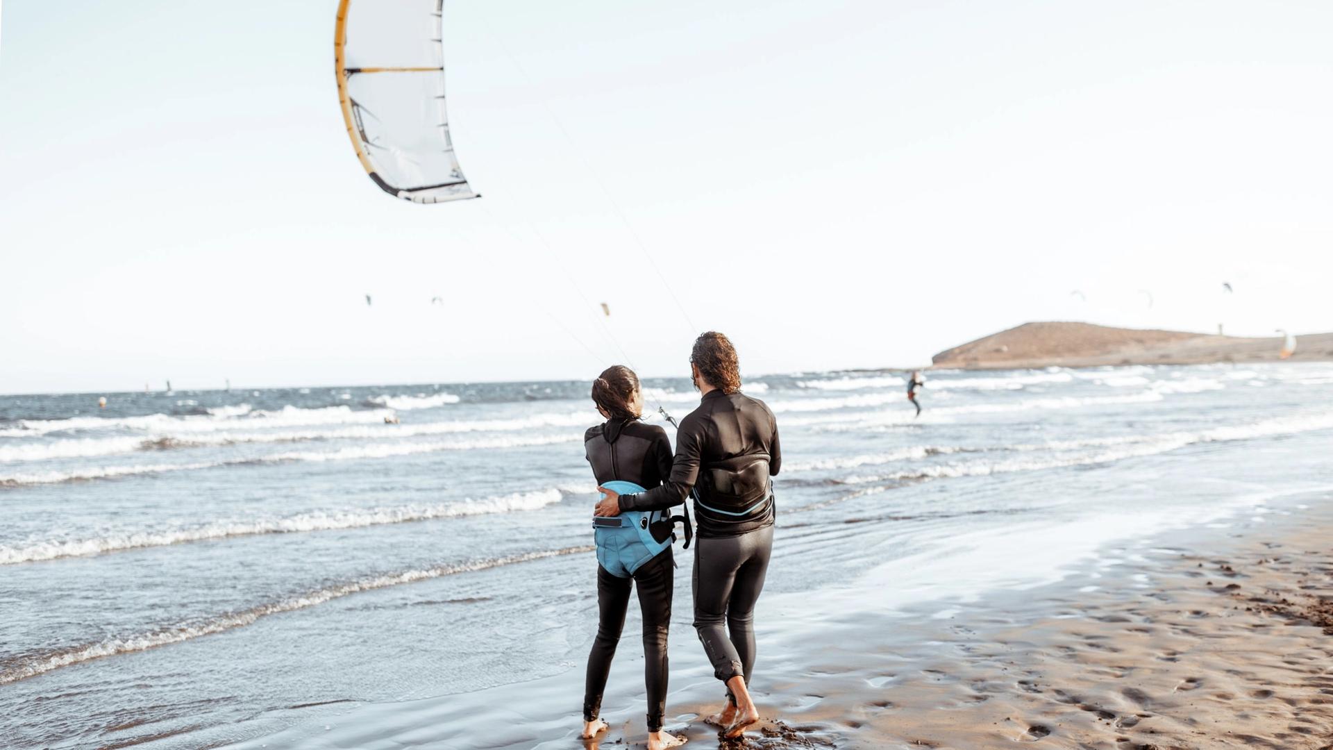 Two people in wetsuits stand on a beach looking out at the ocean where a kiteboarder is in the water and a kite is in the air.