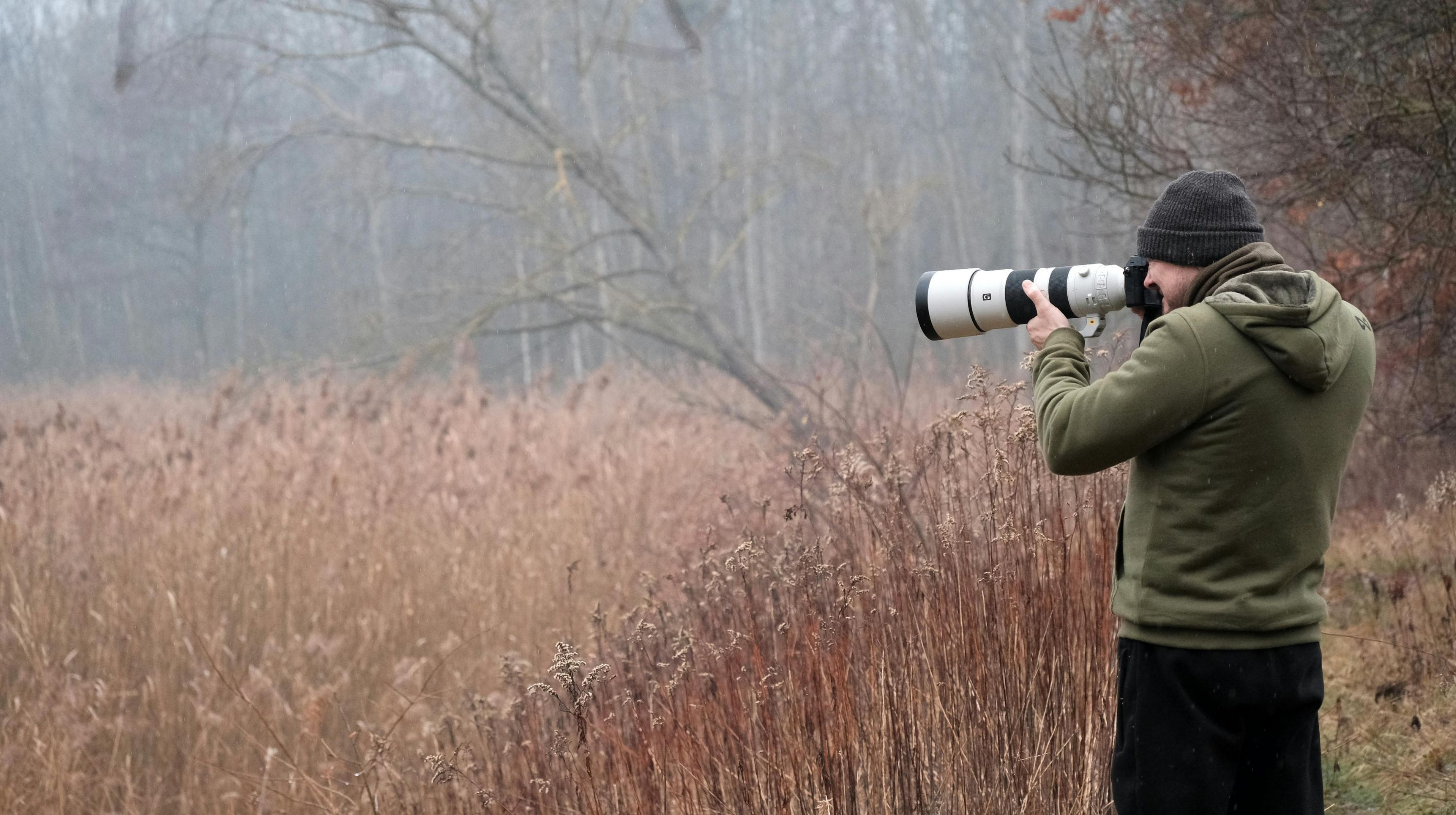 A photographer wearing a green hoodie and a beanie takes a photo with a large telephoto lens in a misty, wooded area with tall, dry grass.