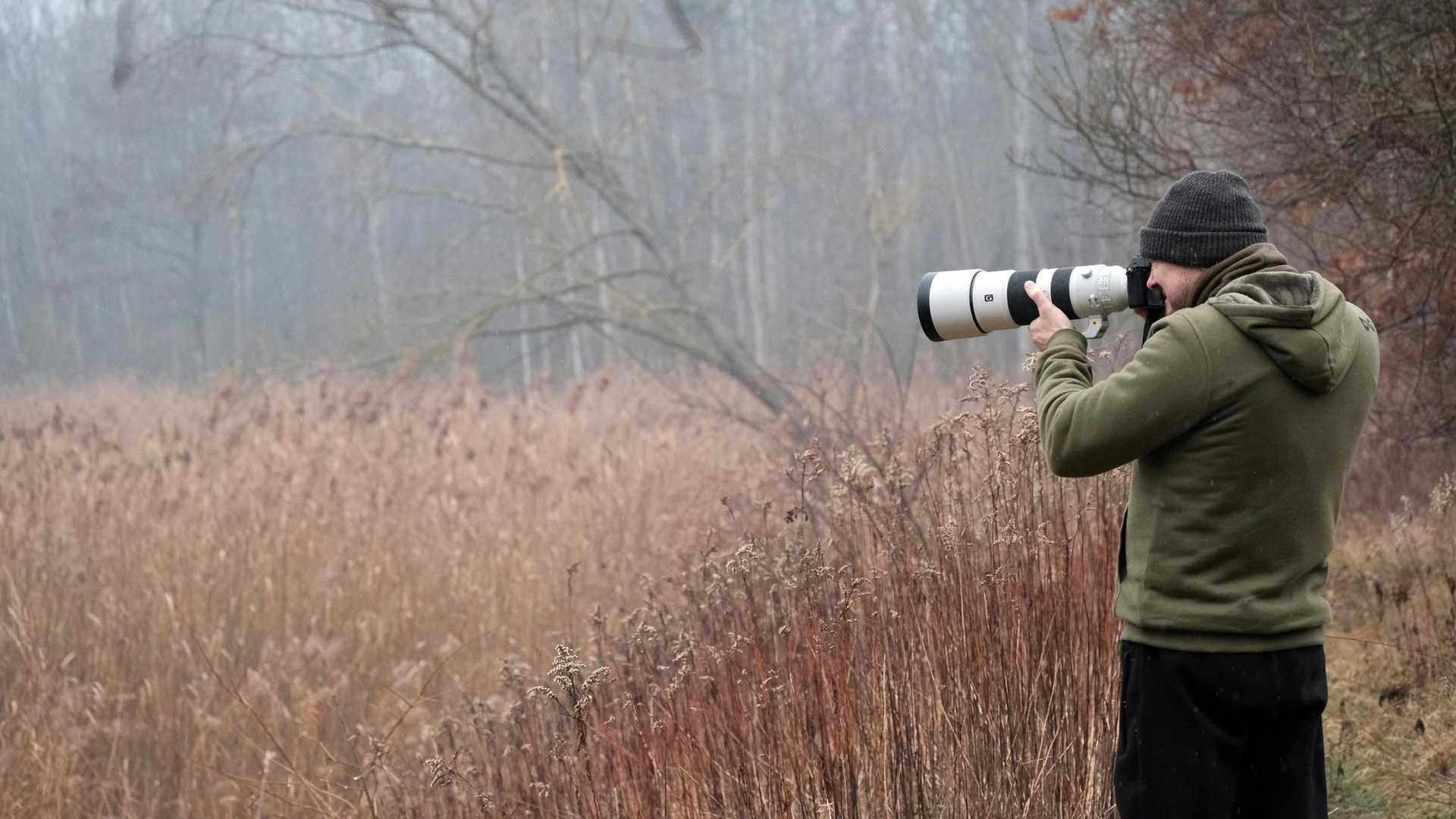 A photographer wearing a green hoodie and a beanie takes a photo with a large telephoto lens in a misty, wooded area with tall, dry grass.